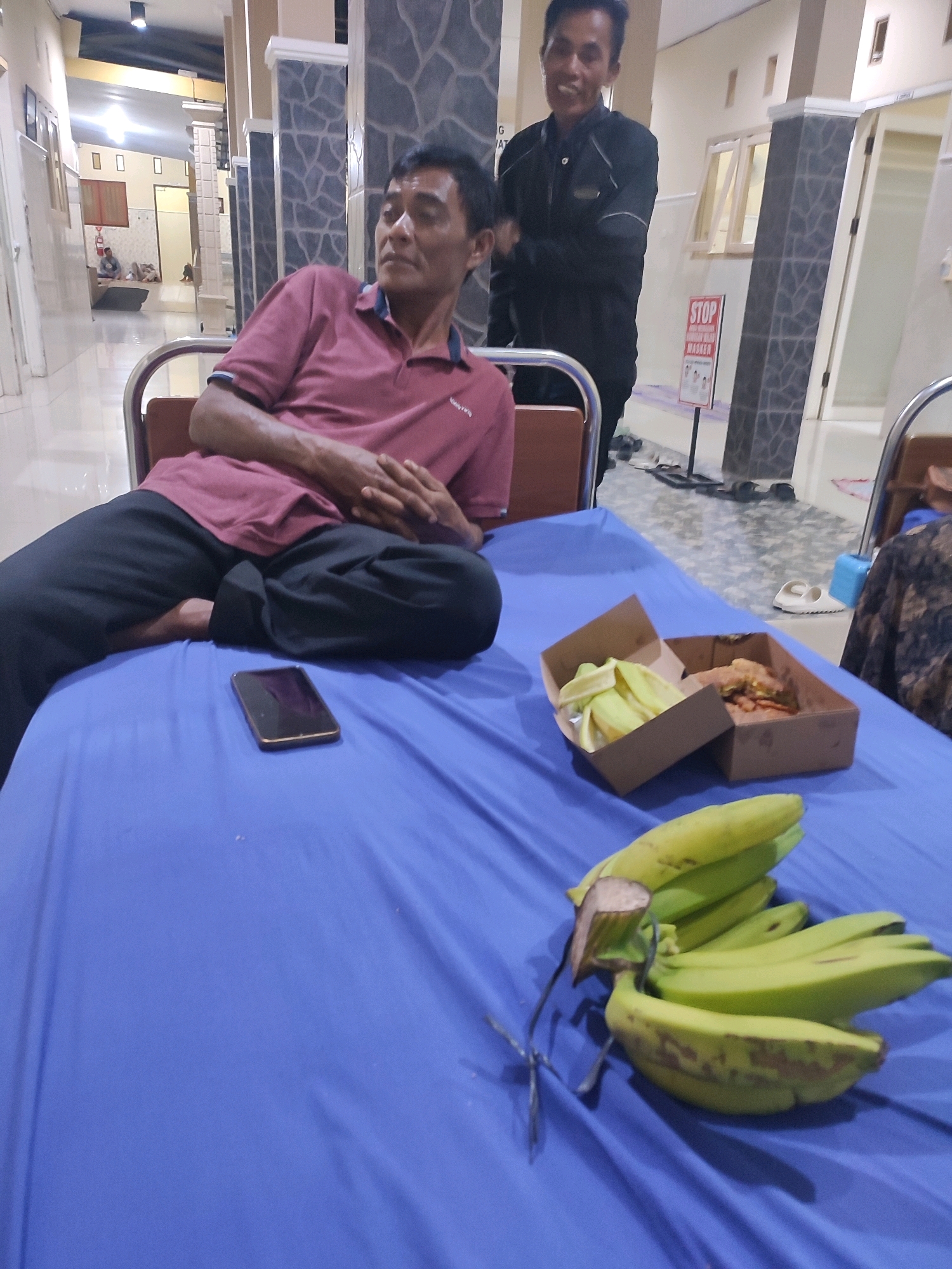 The image depicts a man in a reddish-pink polo shirt and black pants lying on a bed with a blue sheet. His hands are clasped together on his abdomen. To his right, on the bed, is a smartphone and two cardboard containers of food, one of which appears to contain fried chicken and the other peeled bananas. A bunch of green bananas is also placed on the bed in the foreground. In the background, another man in a black jacket stands and smiles. The setting appears to be an indoor facility, possibly a medical clinic or waiting area, with tiled floors, walls, and pillars. A "STOP" sign with Indonesian text and images is visible in the background. The overall lighting suggests an indoor environment. This scene is likely in Sukorejo, Indonesia, as indicated by the context provided.