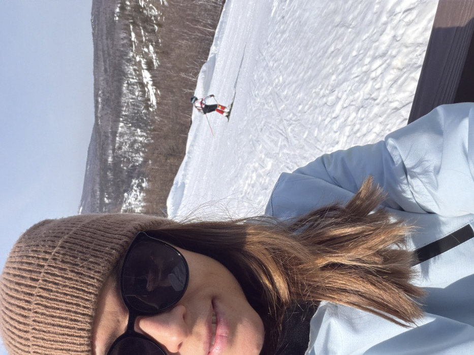 The scene depicts a snowy mountain landscape, identified as Zelenaya Polyana, Russia, under a clear blue sky. In the left foreground, a person is taking a selfie. This individual is wearing a light brown knitted beanie, dark sunglasses, a light blue puffer jacket, and a dark turtleneck; brown hair is visible. A dark railing is present at the bottom right of the frame, suggesting an elevated position. Further into the midground, on a snow-covered ski slope marked with tracks, a second individual is observed actively skiing downhill. This skier wears a white helmet, a white top with red markings, red ski pants, and is holding ski poles. The background features snow-dusted mountains and hills with sparse deciduous and coniferous trees.