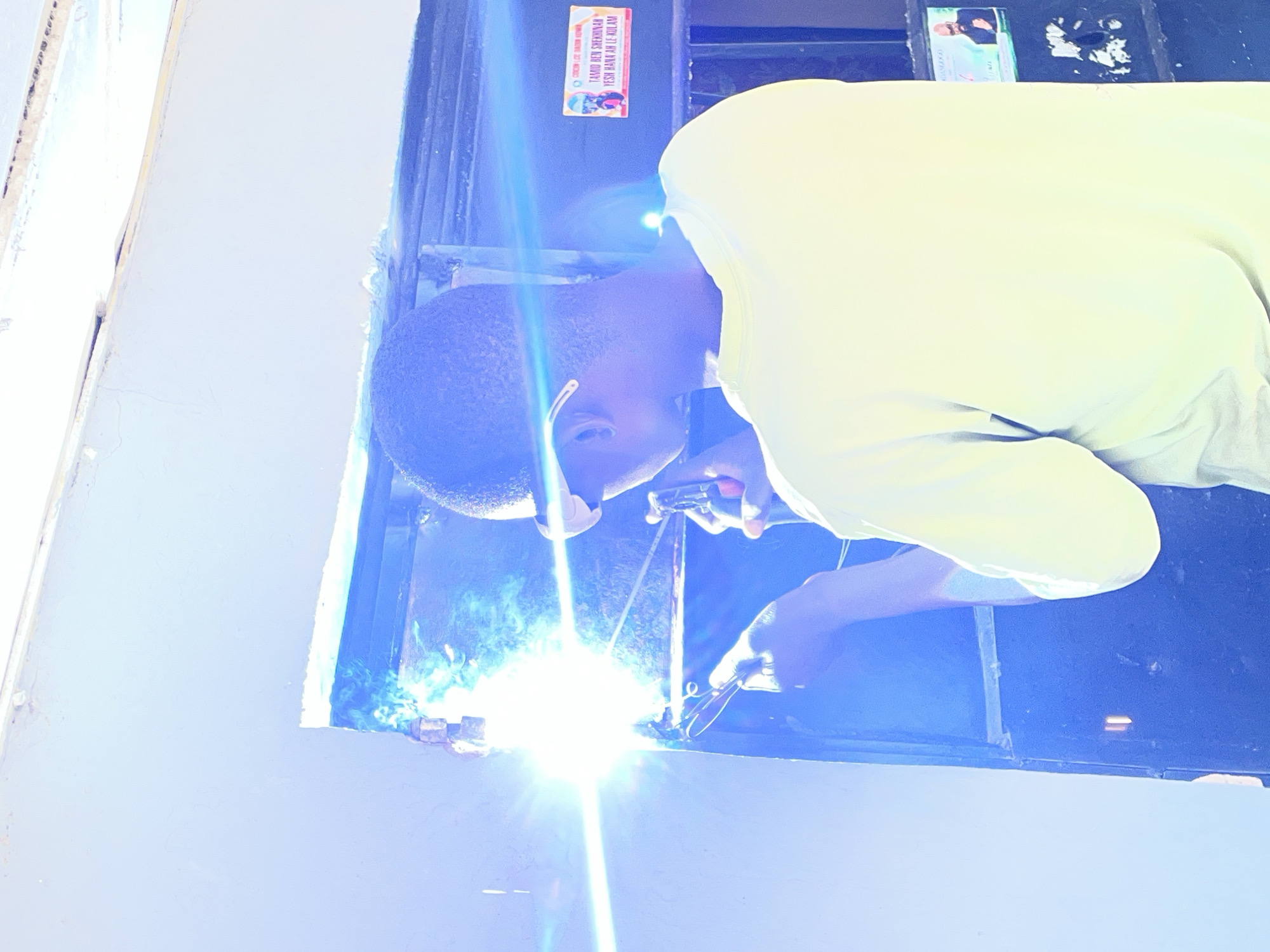 A man wearing a light yellow shirt is welding metal in an open window. The welding torch emits bright light and sparks. A small sticker is on the upper part of the window frame. The location is believed to be Jos, Nigeria.