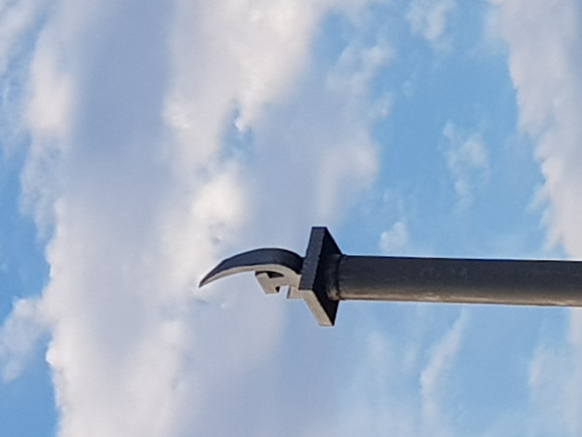 A dark grey, cylindrical pillar topped with a square plinth supports a stylized, metallic sculpture resembling a flame or a sickle against a bright blue sky with scattered white clouds. The sculpture is abstract and metallic, with a curved, upward-sweeping element. The pillar appears to be made of concrete or a similar material. The scene is outdoors. The location is Kurchatov, Kazakhstan, though specific details about the immediate surroundings are not visible.