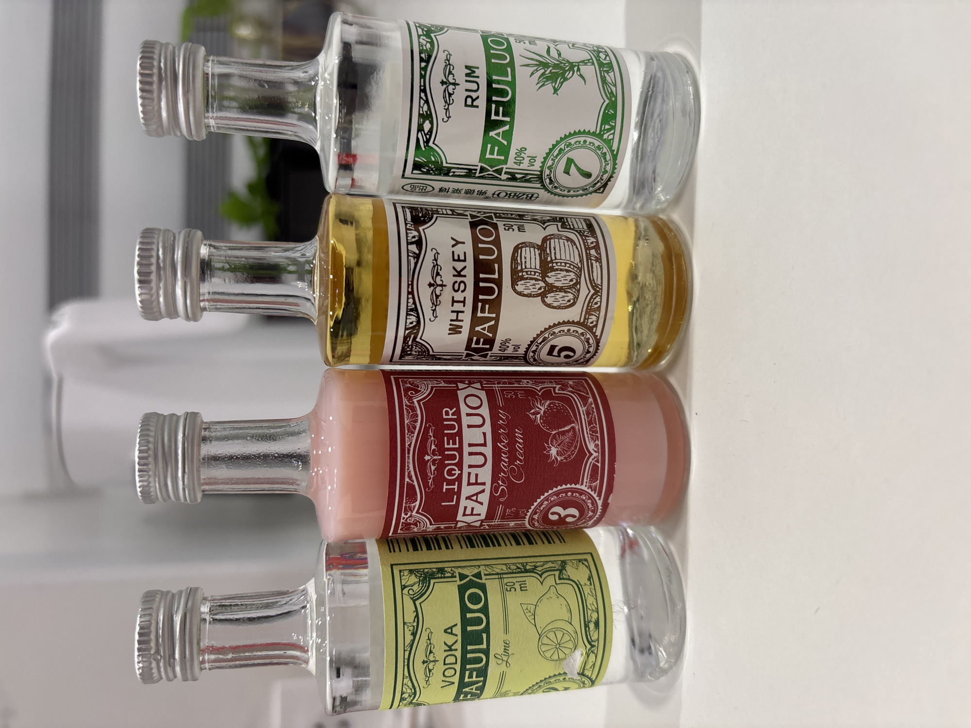The image displays four small glass bottles of alcoholic beverages, arranged horizontally on their sides on a reflective white surface. Each bottle features a silver screw cap and contains a liquid of a distinct color. The background is softly blurred, showing faint green elements, possibly plants.
From left to right (or bottom to top in the original image orientation):
The first bottle holds a clear liquid, identified by its label as "VODKA FAFULUO" with a "Lime" flavor, and a volume of "50 ml".
The second bottle contains a pink, creamy liquid, labeled "LIQUEUR FAFULUO Strawberry Cream". Its label also indicates "17% vol", "50 ml", and the number "3".
The third bottle features a golden-amber liquid. The label reads "WHISKEY FAFULUO", specifying "40% vol", "50 ml", and the number "5". Additional Chinese characters "BZB", "弗德莱", "博", and "出品" are visible.
The fourth bottle, containing a clear liquid, is labeled "RUM FAFULUO", with "40% vol", "50 ml", and the number "7" also printed on its label.
The scene is a static display of these miniature liquor bottles, with no activity taking place. The setting appears bright and clean, possibly indoors, but no specific time of day or location cues are evident beyond the immediate arrangement.