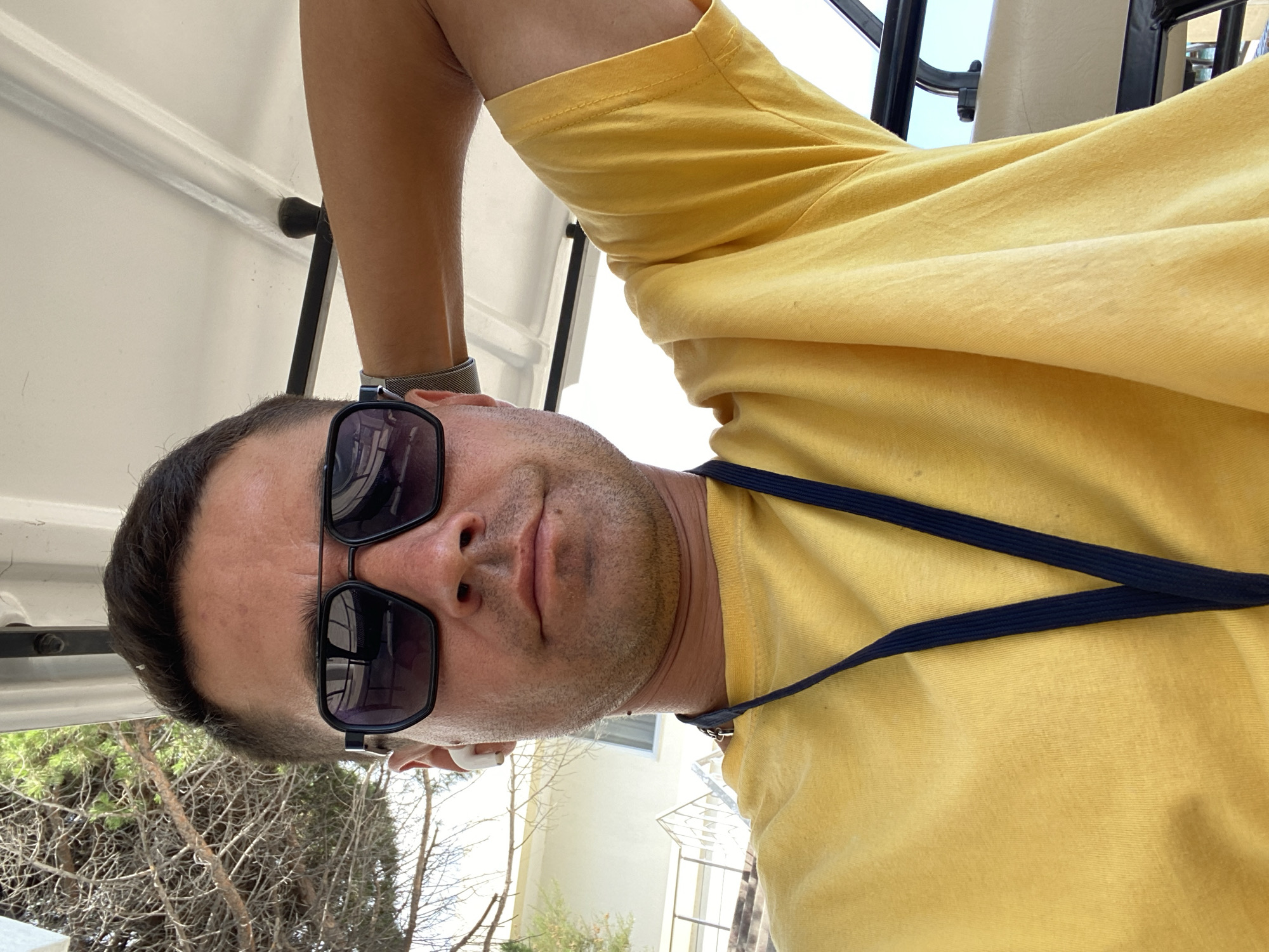 In the city of Koreiz, Ukraine, a man is taking a selfie outdoors, possibly from a golf cart. He is wearing sunglasses, a yellow t-shirt, and wireless earbuds. His arm is resting behind his head. The background shows trees, a building, and stairs.