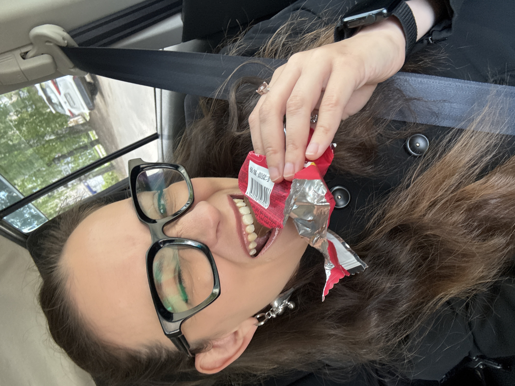 A woman with long brown hair and black-rimmed glasses is in a car, eating a chocolate bar. She is wearing a black jacket and a seatbelt. A silver earring shaped like a teddy bear is visible on her left ear. The background shows trees and another car outside the window. The woman is looking at the camera and winking.
