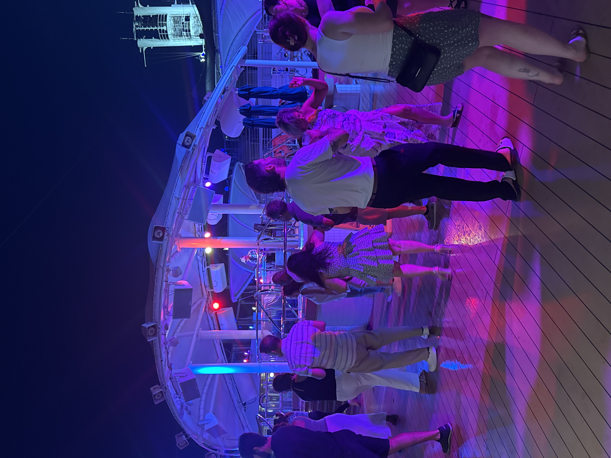 The image depicts a group of people dancing on a wooden deck at night. The scene is illuminated by colorful lights, predominantly purple and blue, casting vibrant reflections on the deck. Some of the individuals are captured mid-dance, with others standing and observing. The background reveals structural elements of what appears to be a ship or a large venue, with lights and railings visible. The overall atmosphere is festive and lively, suggesting an event like a party or a dance gathering. The time of day is clearly night, indicated by the dark sky and artificial lighting.
