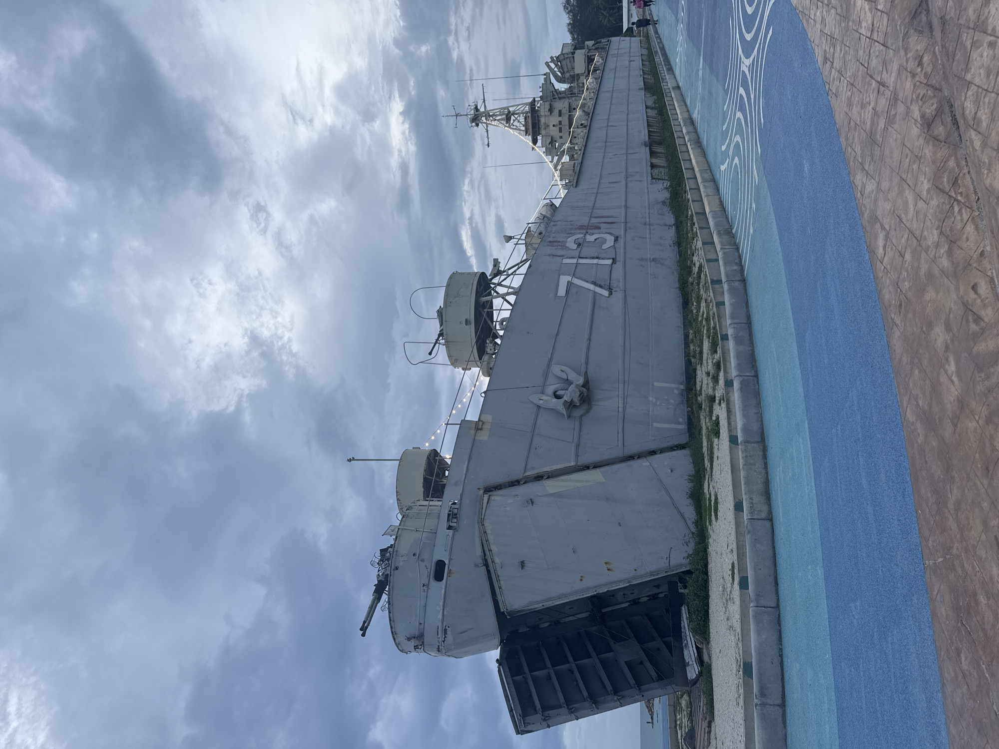 The image shows a naval ship, identified by the number "713" painted on its side, docked along a blue and white patterned waterfront walkway. The ship appears to be an amphibious transport dock with its stern gate open. A small group of people can be seen in the background near the ship's upper deck. The sky is cloudy. This scene depicts the HTMS Pangan naval ship, a museum piece and landmark in Thong Sala, Thailand.