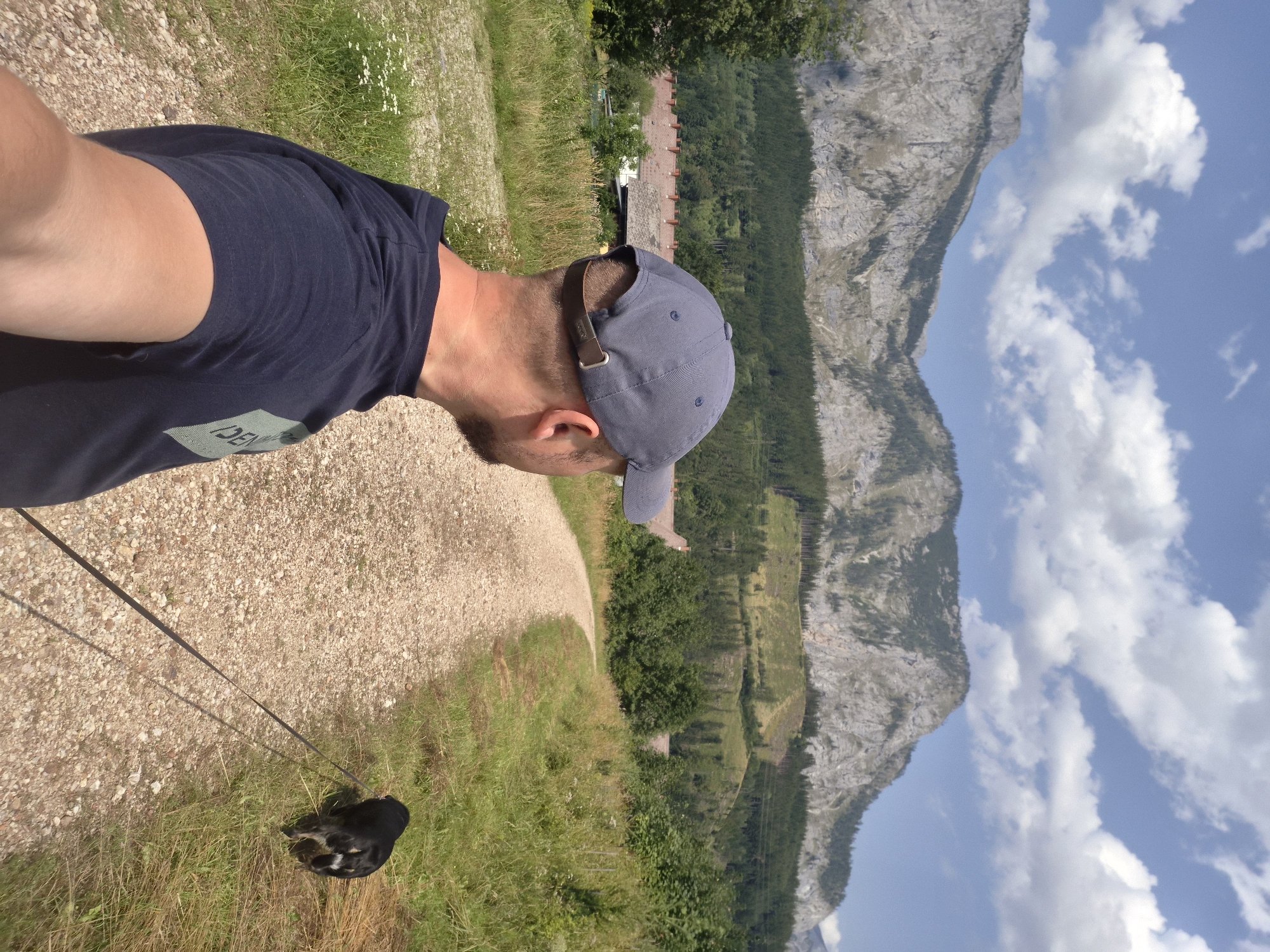 A man wearing a dark blue t-shirt and a grey baseball cap is standing on a gravel path, facing away from the camera. He is holding a black dog on a leash. The dog is walking ahead of him on the right side of the path. The path curves to the left and goes into a wooded area. In the background, there are mountains with some trees and buildings visible. The sky is blue with some white clouds. The scene appears to be a rural area in Eisenerz, Austria, possibly near a hiking trail.