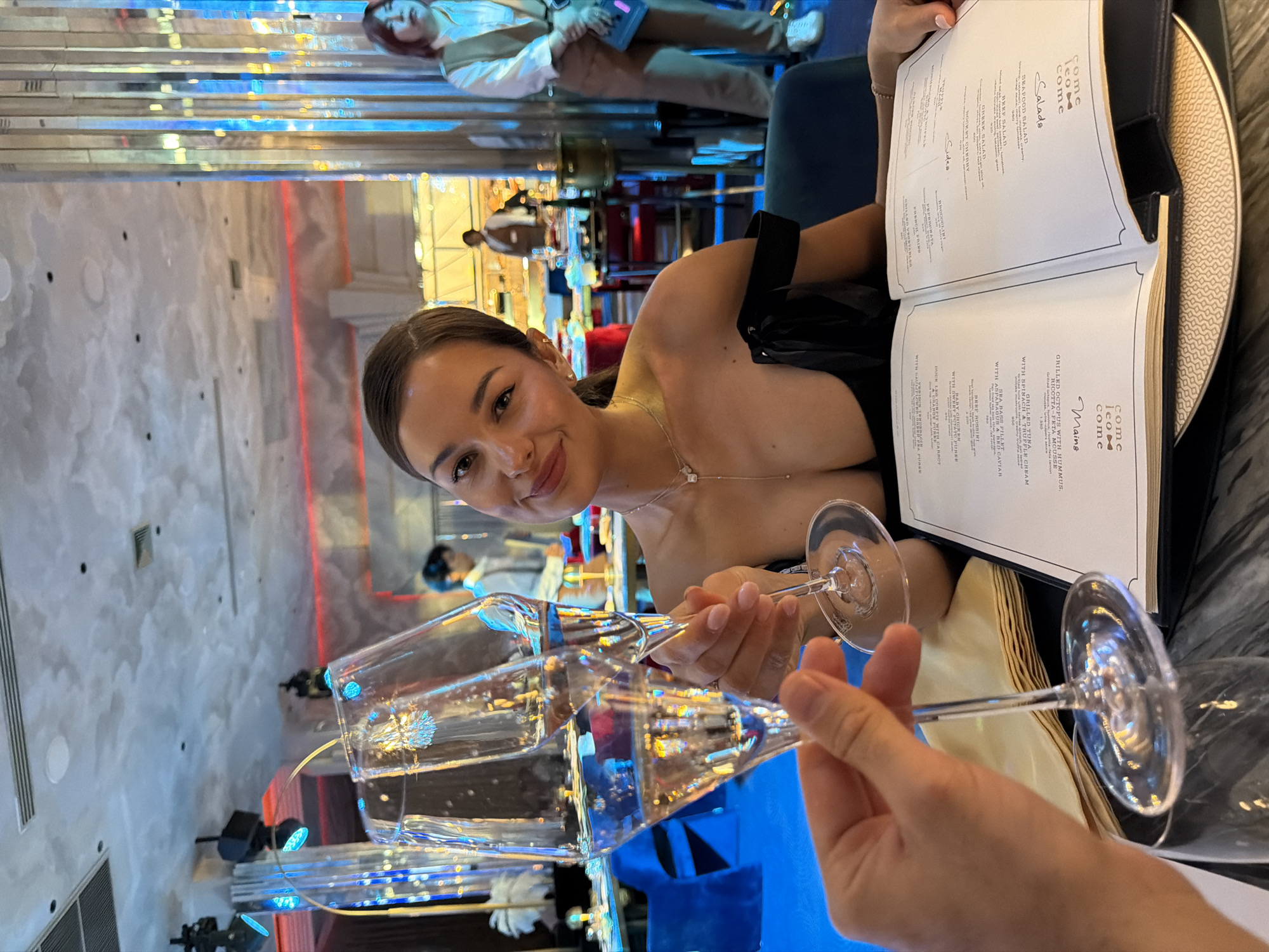 A young woman sits at a table in what appears to be an upscale restaurant. She has fair skin, brown hair pulled back, and is wearing a low-cut black top with a delicate necklace. She smiles at the camera while holding a champagne flute, presumably filled with champagne, as another person's hand enters the frame with a second flute, suggesting a toast.
On the table, there's a large, open menu with the restaurant name "Come Teo Come" visible. The menu lists various dishes like seafood salad, beef salad, and grilled octopus.
The restaurant appears to be modern, with a mirrored wall and lighting fixtures, with an interior design possibly in Choeng Thale, Thailand. Other patrons are visible in the background.