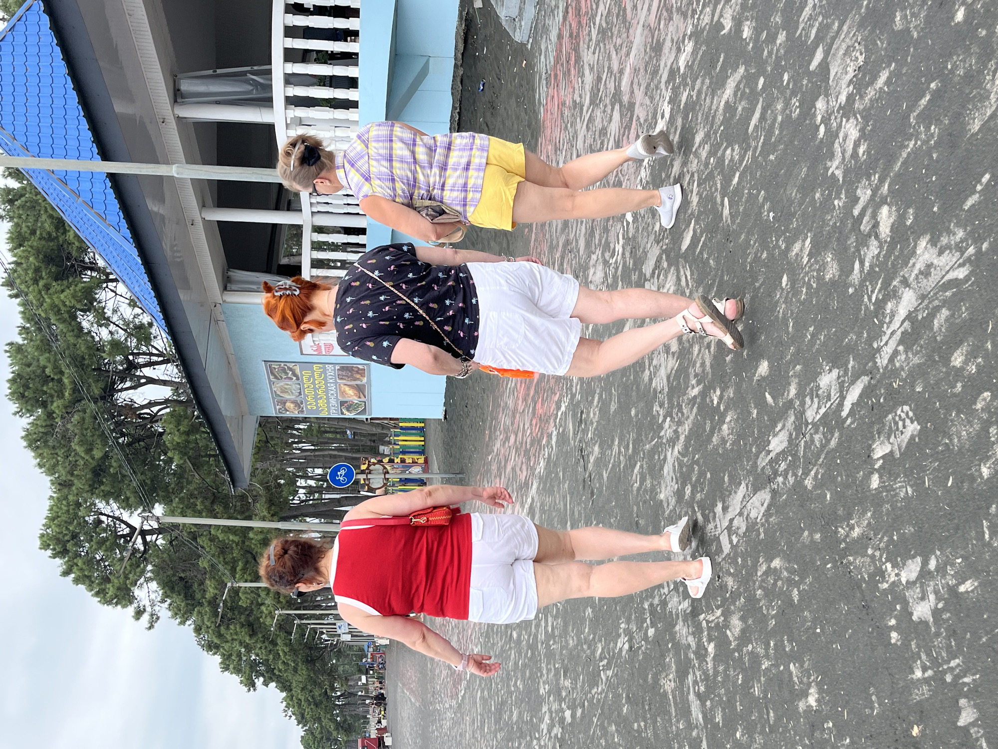 Three women are walking away from the camera on a black sandy surface, which appears to be a promenade or beach area in Ureki, Georgia. The woman in the foreground is wearing a red tank top and white shorts. The middle woman is wearing a black t-shirt with a flamingo print and white shorts. The woman on the right is wearing a plaid shirt and yellow shorts. Behind the women, there is a light blue building with a blue tiled roof and a white railing. A line of pine trees runs along the background. The sky is overcast.
