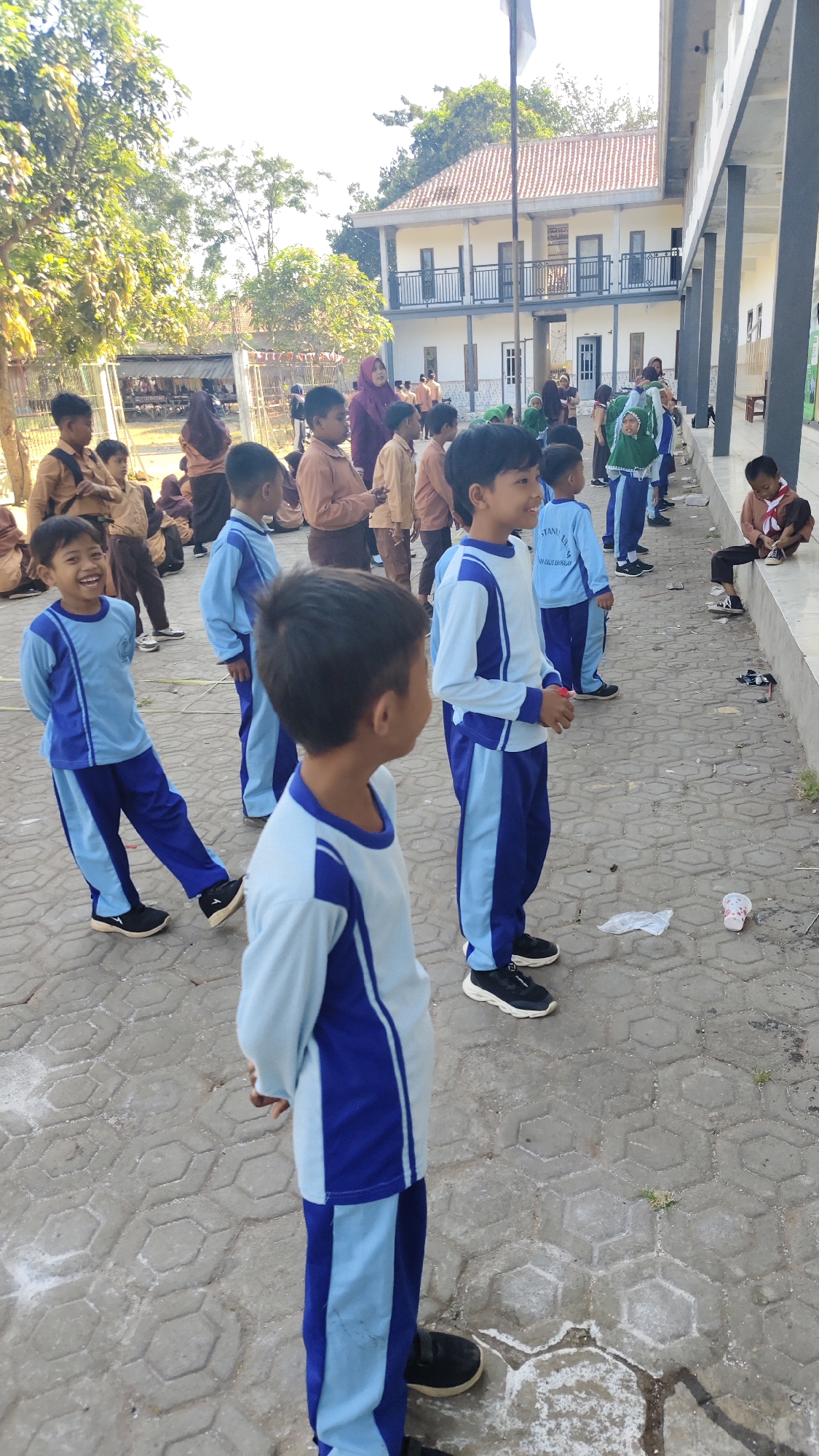 In Banyubunih, Indonesia, an outdoor school scene unfolds under a bright sky. A group of elementary school children, all wearing light blue and navy blue uniforms, are gathered on a paved courtyard. Some are standing in formation, while others are interacting. In the foreground, a boy in a light blue shirt and navy blue pants grins broadly, his left leg extended. To his right, another boy in a similar uniform stands facing away from the camera. Further back, more children in uniform are visible, some in a line, and a few more are seated or standing casually. A two-story building with white walls and a tiled roof, featuring balconies and windows, forms the backdrop, suggesting a school or educational institution. Trees and foliage are visible around the perimeter of the school grounds. The pavement is made of hexagonal interlocking concrete blocks, with some debris and white powder scattered on the ground.