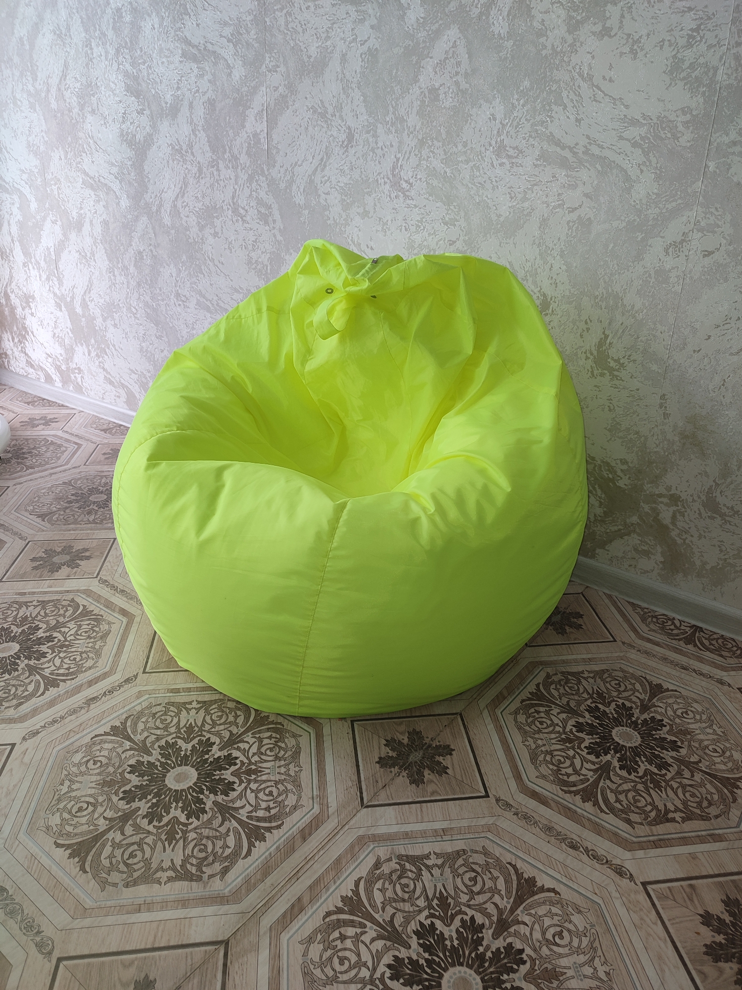 A bright yellow beanbag chair sits on a patterned tile floor in a room with textured walls. The beanbag chair is round and appears to be made of a synthetic material. The floor tiles are octagonal with ornate brown and beige patterns. The walls are a light color with a textured finish that resembles stucco or wallpaper. The image is taken from a medium distance, showing the beanbag chair prominently in the center of the frame. There are no people or other objects visible in the scene.