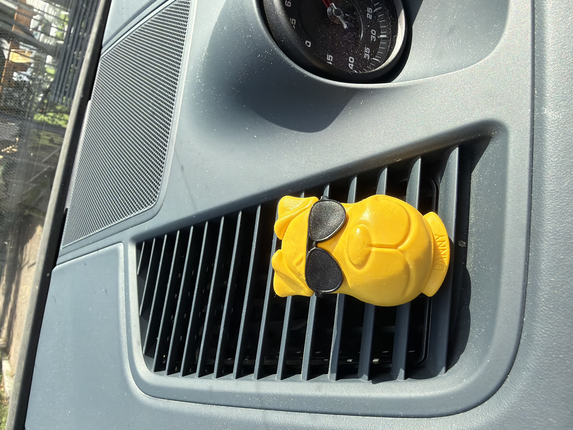 A yellow dog-shaped air freshener with black sunglasses is clipped to the horizontal slats of a car's air vent. The car's dashboard is dark gray and dusty. To the right of the vent is a black gauge with white markings, appearing to be a speedometer, with a red needle pointing towards 50. The background is blurred, showing sunlight filtering through trees and a fence, suggesting an outdoor setting. The car is parked in an outdoor environment. The image was taken on Rustaveli Avenue in Tbilisi, Georgia.