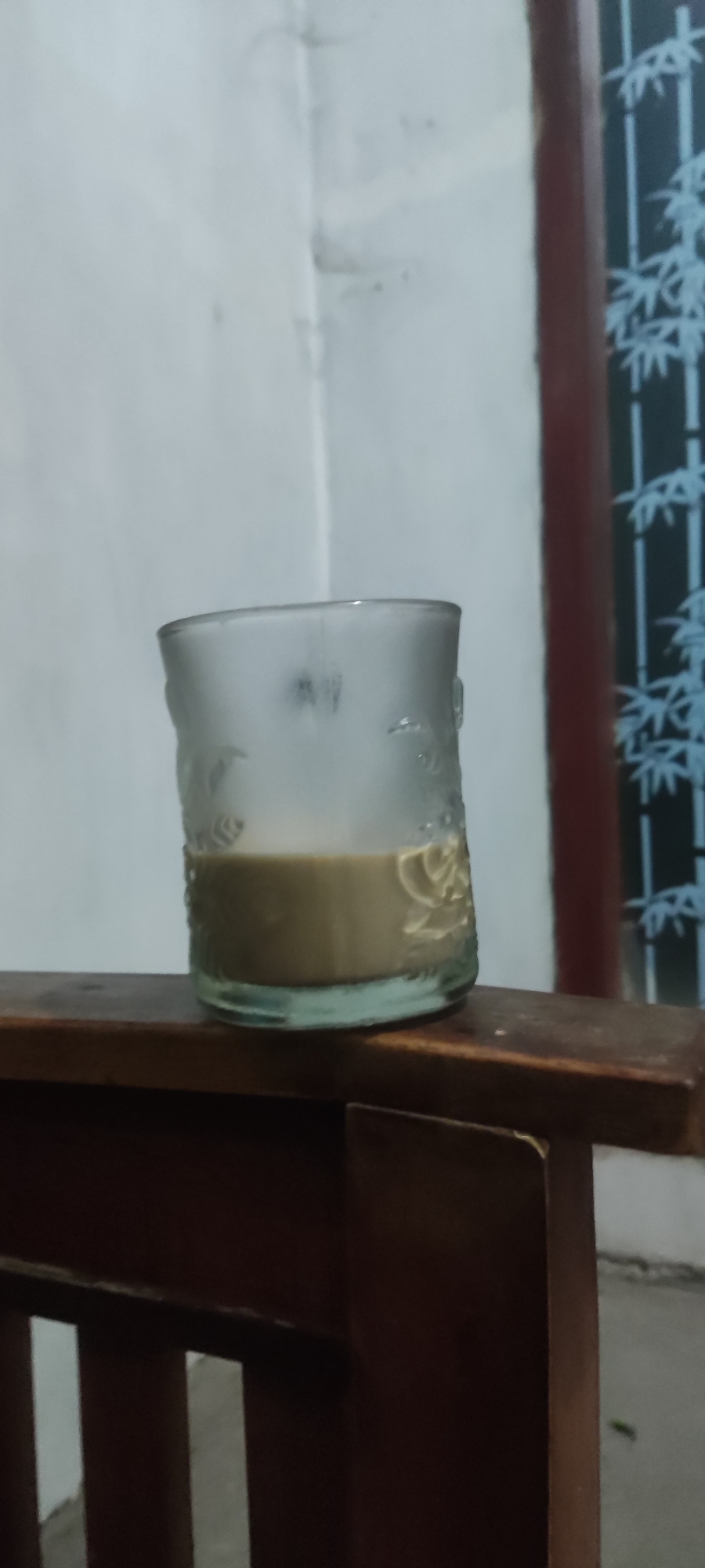 A glass of light brown liquid, possibly coffee or tea, sits on a wooden surface. The glass is partially frosted and has a textured pattern around its exterior. Behind the glass, a wall is visible, which appears to be a light color and possibly textured or slightly damaged. To the right of the wall is a dark-colored vertical element, possibly a doorframe or window frame, with a light-colored decorative pattern of bamboo stalks. The wooden surface appears to be part of a piece of furniture, likely a chair or bench, with visible slats. The overall lighting suggests it might be indoors or in a shaded outdoor area, possibly in the city of Karangwangun, Indonesia, given the context. There are no people visible in the image.