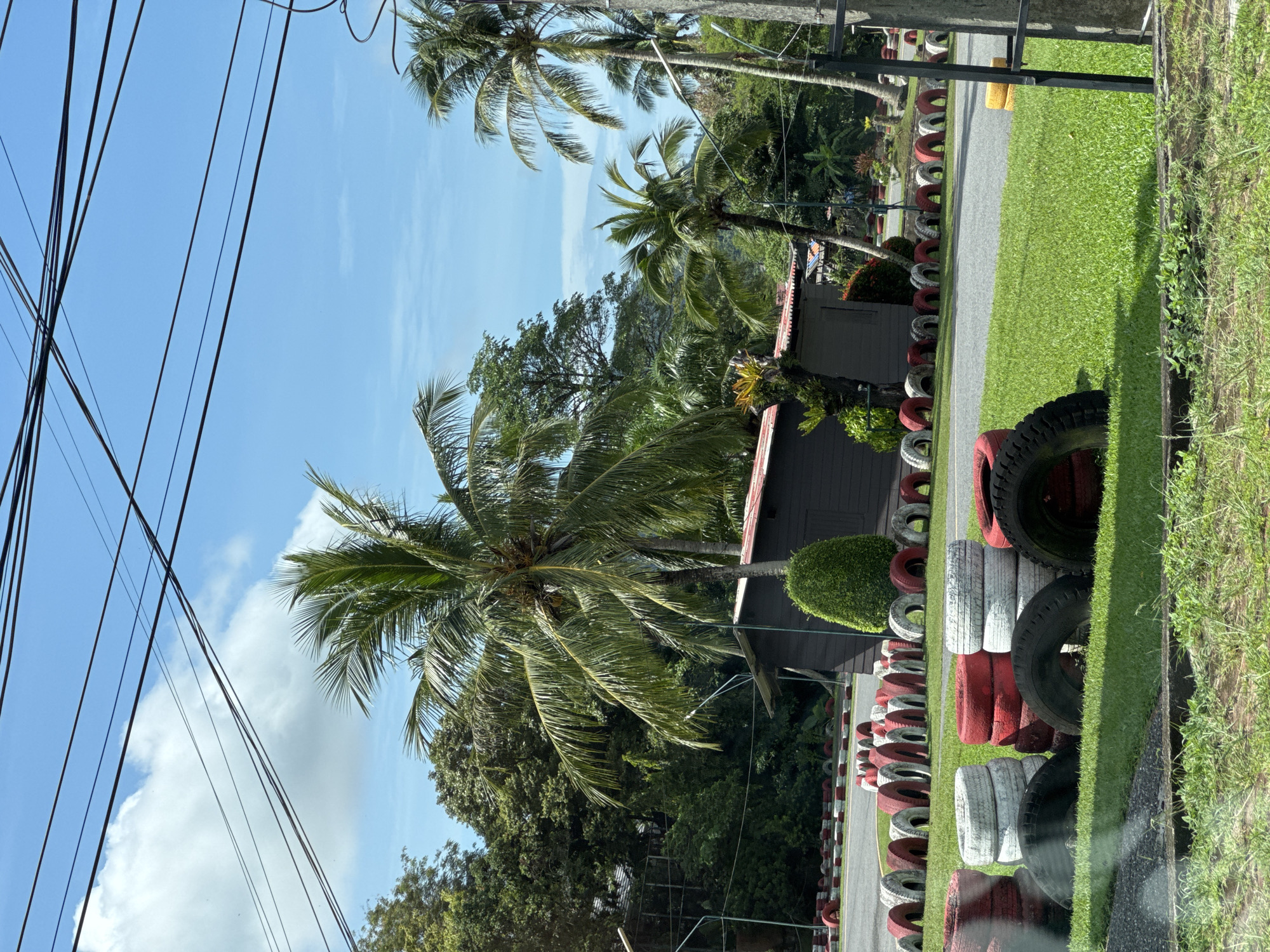 The image depicts an outdoor scene in Kathu, Thailand, likely a go-kart track or a similar recreational facility. The foreground features several stacks of tires, some painted red and white, others black. These tires appear to be serving as safety barriers. Beyond the tires, a paved track is visible, also lined with red and white tires. In the middle ground, a dark wooden structure with a red roof stands amidst lush green vegetation, including a large coconut palm tree directly in front of it. Several other palm trees and dense foliage are present in the background. Overhead, a network of power lines crisscrosses a bright blue sky dotted with scattered white clouds. The overall impression is of a tropical, sunny day at an outdoor entertainment venue. There are no people or animals visible in the image.