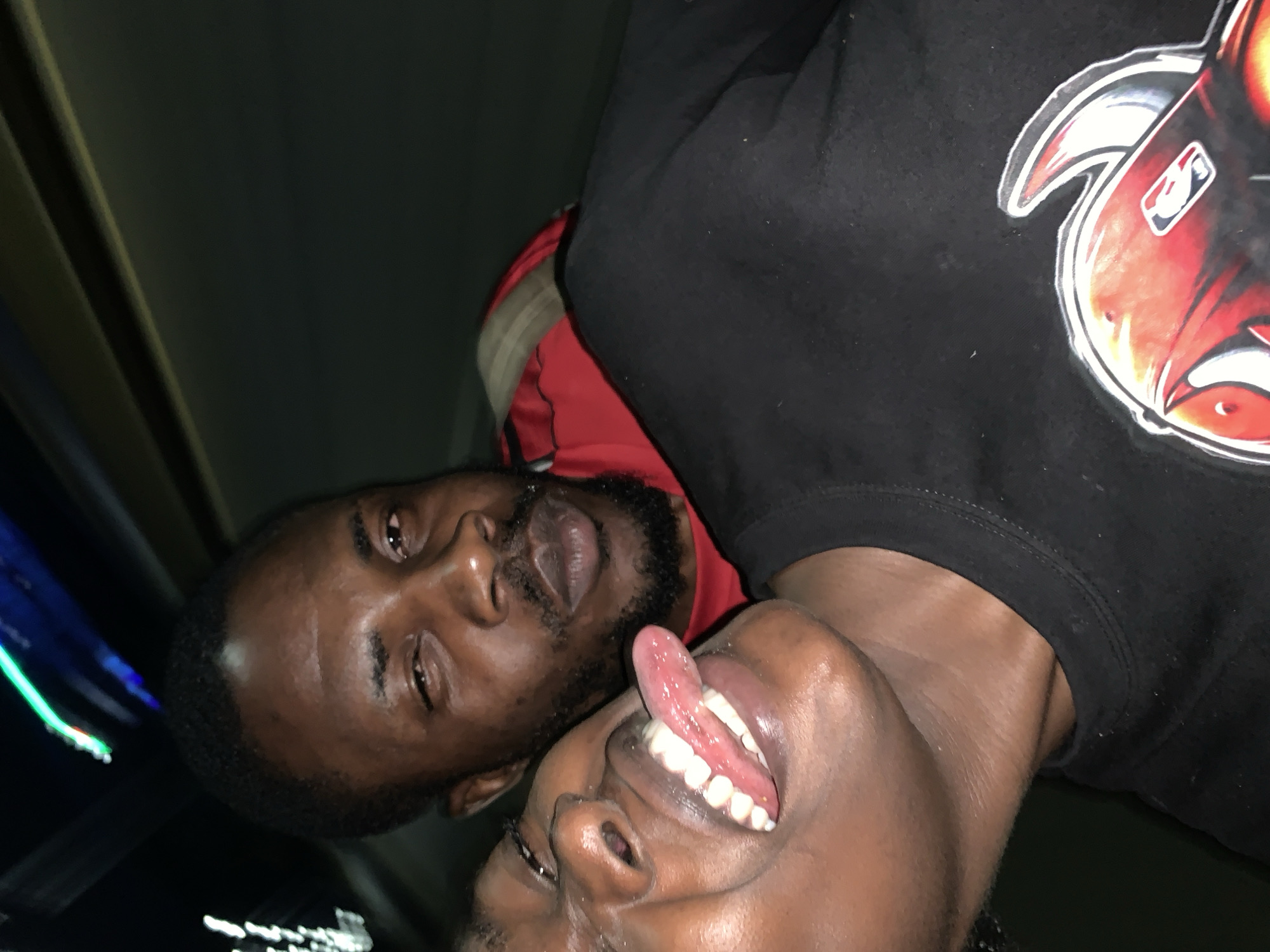 Two dark-skinned males are visible. The male on top wears a red hoodie under a black sweatshirt with an NBA logo. The male on the bottom has his mouth open with his tongue sticking out. The location is presumably the city of New Stadium, Nigeria. The image is taken in a dark, low-light environment.