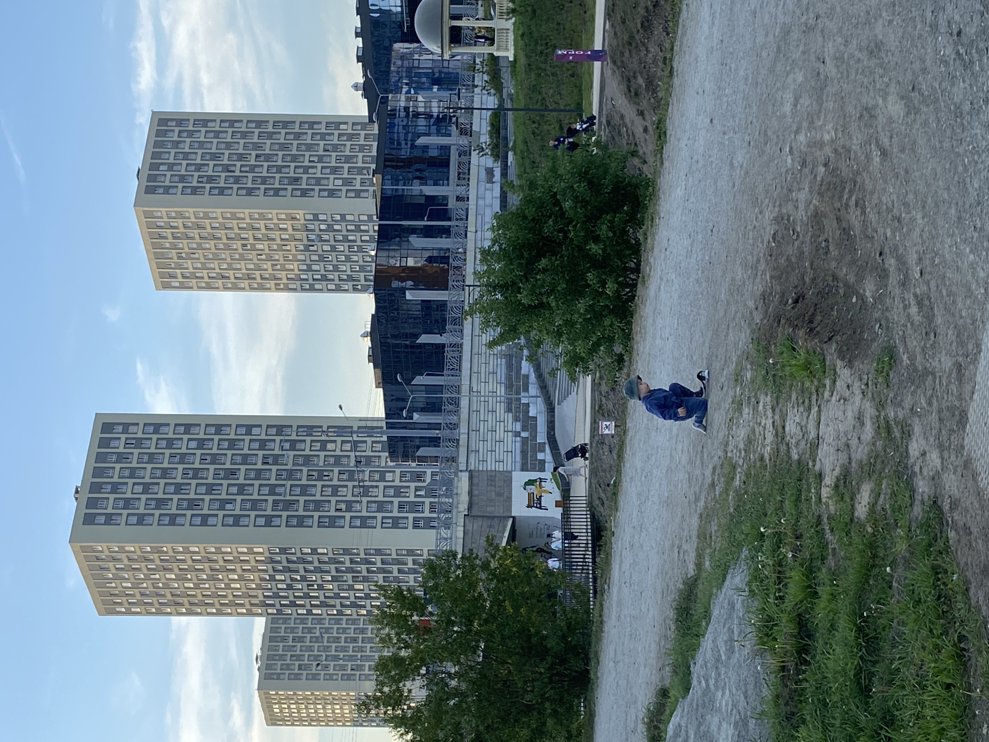 The image shows a view of a park in Yekaterinburg, Russia, with modern buildings in the background. There are three tall buildings with repetitive window patterns visible. A walkway or path runs through the foreground, where a child in a blue outfit sits. There are trees and greenery along the path. In the background, there's a structure resembling a gazebo or pavilion.