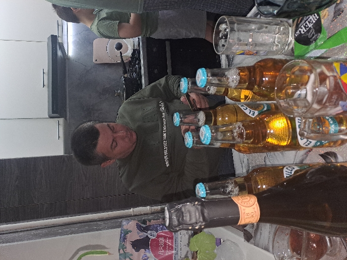 A kitchen counter is cluttered with empty beer bottles and glasses. A man in a green sweater stands by the counter, apparently having consumed some of the beer. Another person is visible in the background. The scene suggests a party or gathering.