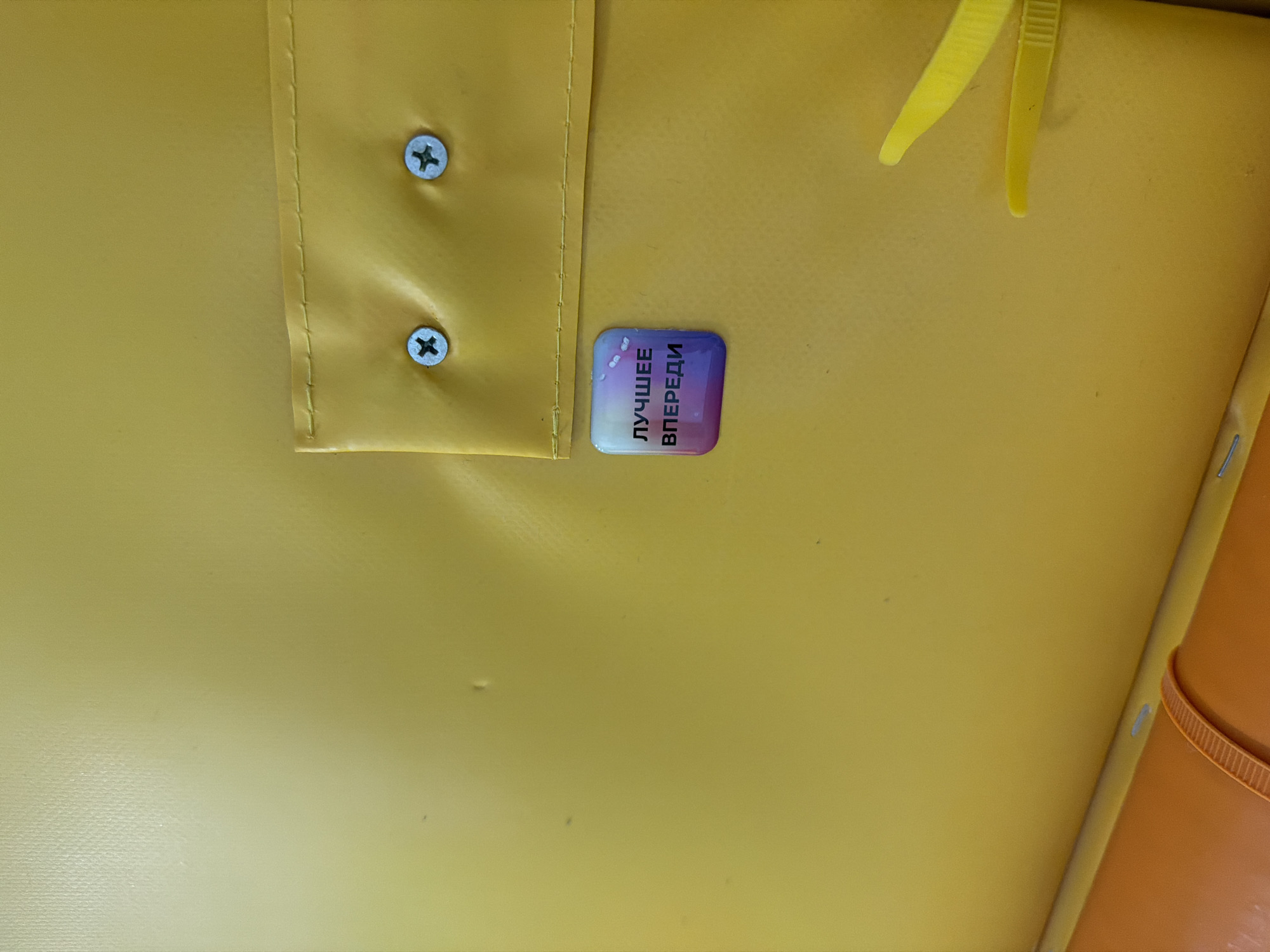 The image shows a close-up of a yellow surface, likely a padded interior. Two metal screws fasten a section of the material that appears to be folded over, with a visible seam. Adjacent to this is a small, rectangular sticker displaying text in Russian: "ЛУЧШЕЕ ВПЕРЕДИ" (meaning "The best is yet to come"). A yellow plastic object, possibly a fastener or clip, is partially visible at the top right. A section of what may be an orange container or object is visible on the lower right side of the photo. The location of Chaikovsky, Russia, is not apparent from the visual information provided.