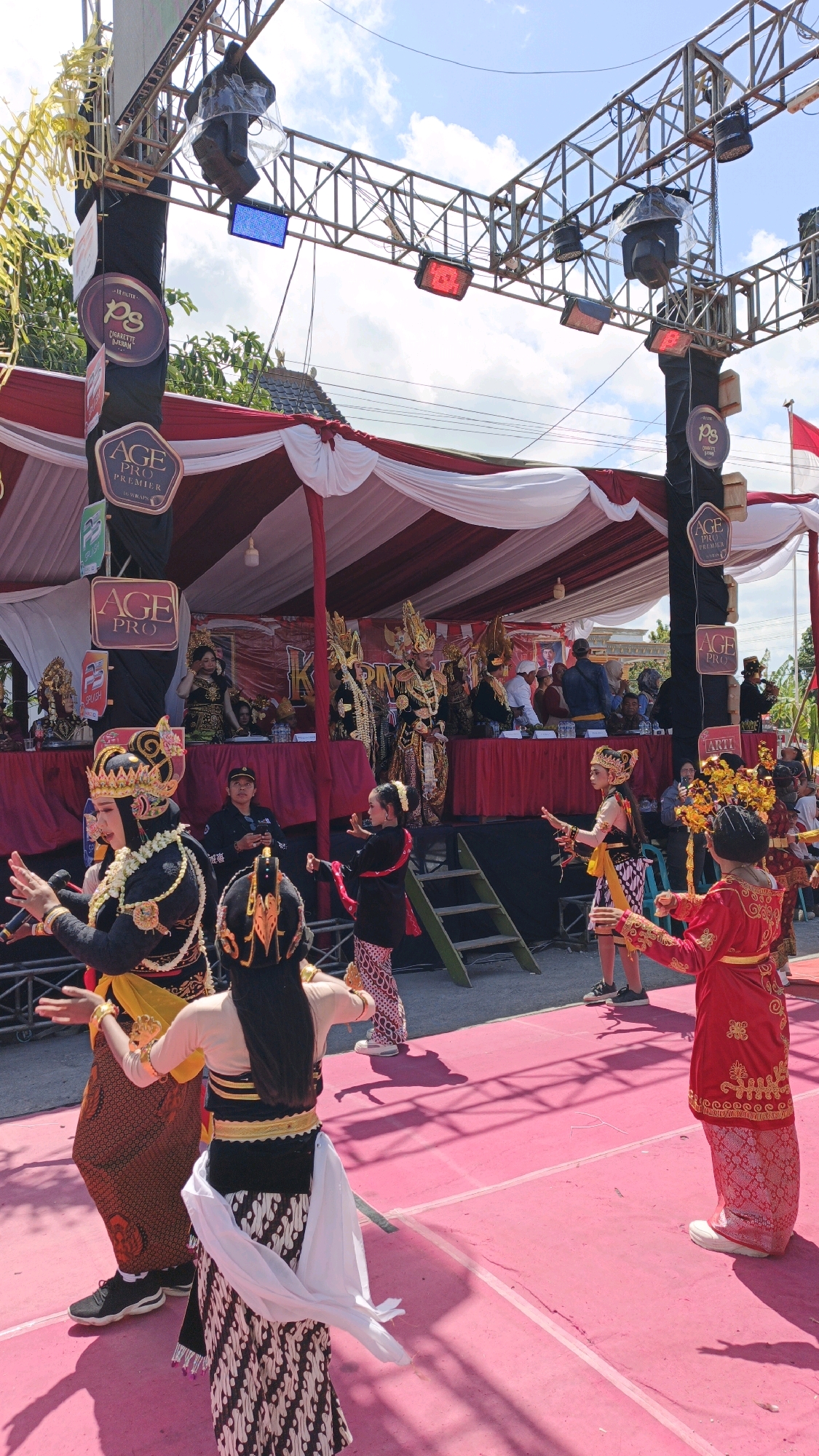 In Gesikan, Indonesia, a vibrant cultural performance unfolds outdoors under a bright, partly cloudy sky. A stage, adorned with a red and white striped canopy, serves as the backdrop for performers dressed in traditional, ornate attire. Several individuals, likely dancers, are actively engaged in a performance on a bright pink carpeted area in the foreground. One performer, wearing a red and gold costume, faces away from the camera, while another, in a black and gold outfit, is positioned to the left, seemingly mid-dance with arms extended. The stage itself features multiple people seated behind a table, observing the performance. In the background, a large metal truss structure supports various lighting equipment. Several banners with logos are visible, indicating sponsorship. The overall atmosphere suggests a festive event or cultural gathering.
