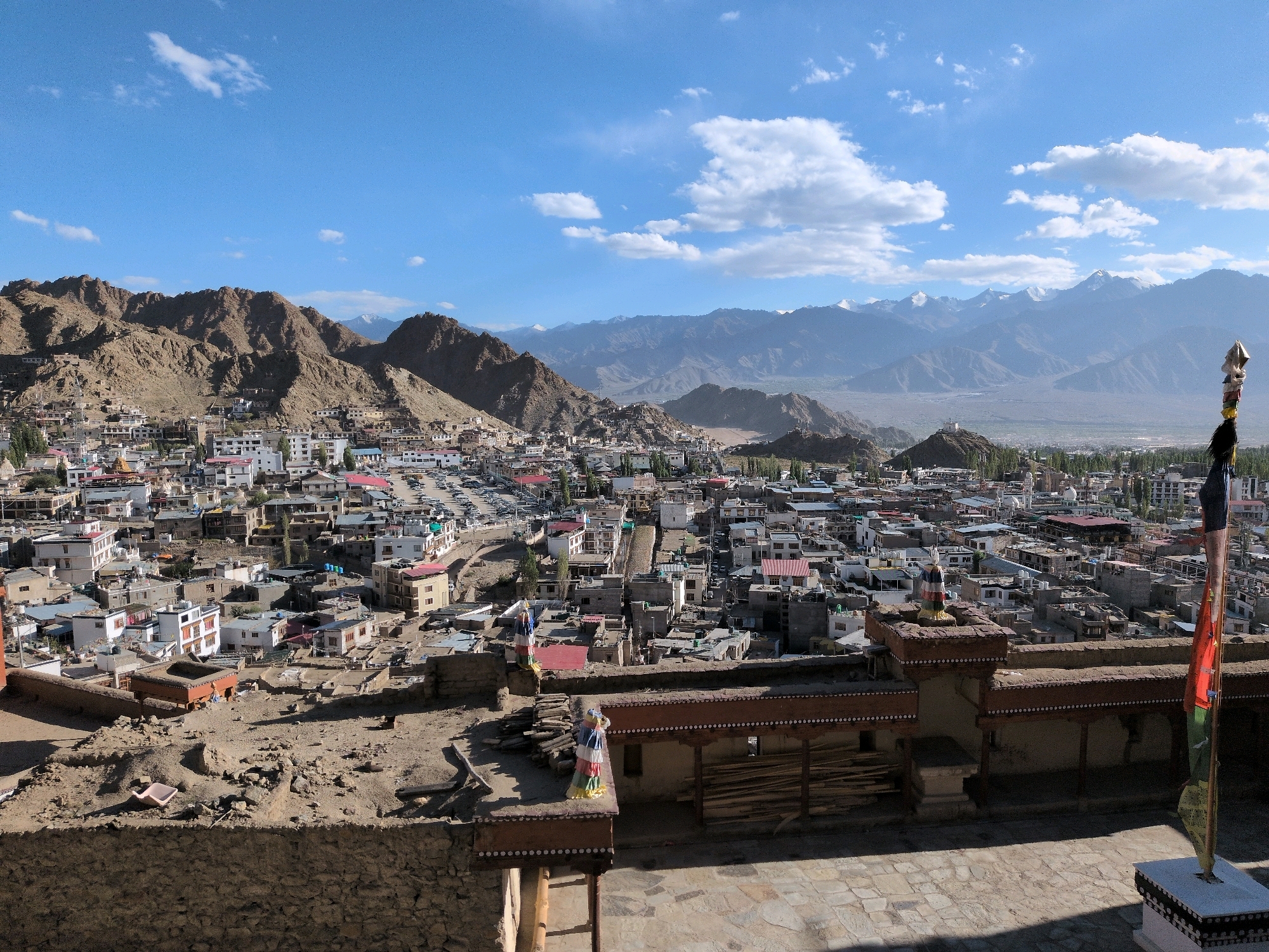 This is a wide, aerial view of Leh, India, captured on a sunny day with a bright blue sky dotted with scattered white clouds. The city, nestled in a valley, is characterized by its densely packed buildings with white and red roofs, spread across the foothills of a large, rugged mountain range. The architecture appears traditional, with many multi-story structures. In the foreground, there is a stone wall and a paved courtyard, with a prayer flagstaff prominently displayed on the right, featuring colorful prayer flags. Further into the city, a mosque's white dome is visible, alongside other buildings and what appears to be a parking lot filled with cars. Beyond the city, the dramatic mountain landscape stretches into the distance, with snow-capped peaks visible on the horizon. The scene is devoid of any visible people.