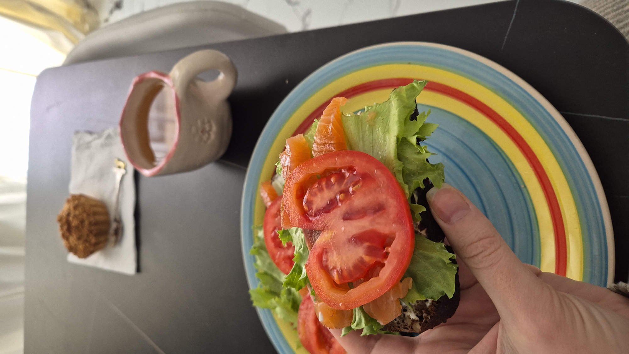 On a black surface, a colorful plate sits beside a cream-colored mug with a pink rim, a white napkin, a spoon, and a muffin. The plate features concentric circles of light blue, yellow, and red. A hand holds a sandwich on the plate, featuring dark bread topped with green lettuce, orange salmon, and red tomato slices. The scene is likely an indoor setting. There is no indication of Batumi, Georgia.