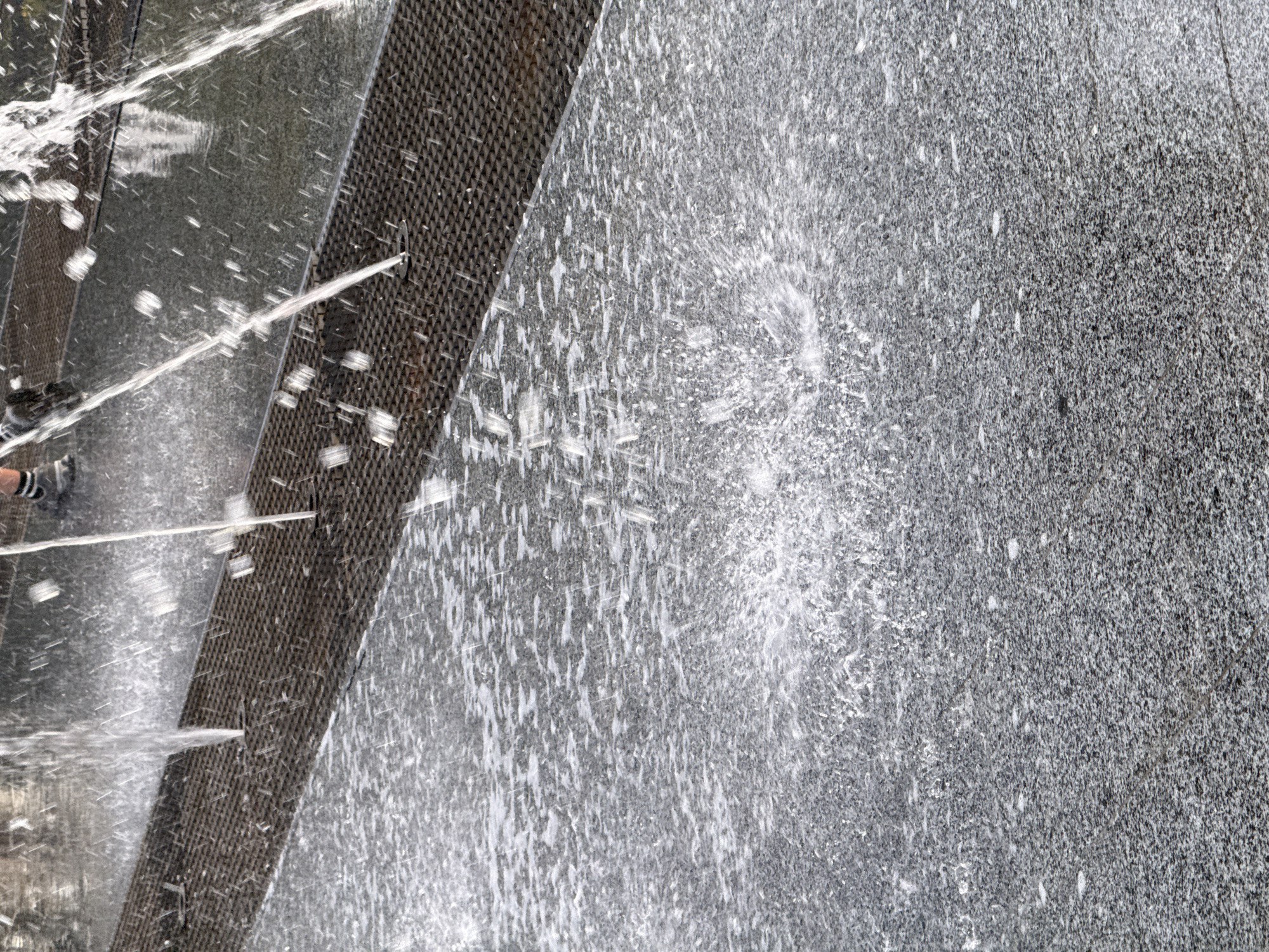 This media file depicts a close-up view of a water feature, likely a fountain, in Piatykhatky, Ukraine. The primary subjects are water and the textured surfaces it interacts with. The scene is dominated by a diagonal band of dark, metallic grating that runs across the left side of the frame, with water spraying from it and onto a speckled, gray stone surface on the right. The stone appears to be granite, characterized by its granular texture and variations in gray tones.
The activity taking place is the spraying of water. Streams and droplets of water are visible in various states of motion – some are sharp, jet-like streams emanating from the grating, while others are dispersed into a fine mist or are captured as splashes and droplets on the surfaces. The water creates a dynamic and somewhat abstract pattern against the solid backgrounds.
Notable details include the contrast between the uniform gray of the stone and the dark, geometric pattern of the grating. The play of light on the water causes some areas to appear bright white, highlighting the movement and texture of the spray. The time of day and weather are not explicitly indicated, but the bright lighting suggests it is daytime with clear conditions. No emotions are discernible as the subjects are inanimate. There is no visible text in the image. The context of Piatykhatky, Ukraine, provides a geographical reference for the location of this water feature.