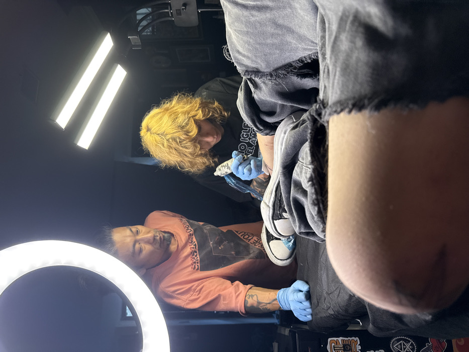 In a tattoo studio, a man with dark hair and a coral shirt lies on a table, getting a tattoo on his foot. The tattoo artist, with blonde curly hair, wears gloves and a black shirt, focused on the process. Artificial light sources surround them: a ring light near the man and two linear lights above the artist. Distressed grey denim shorts are in the foreground. Stickers are visible on the table near the man's feet.