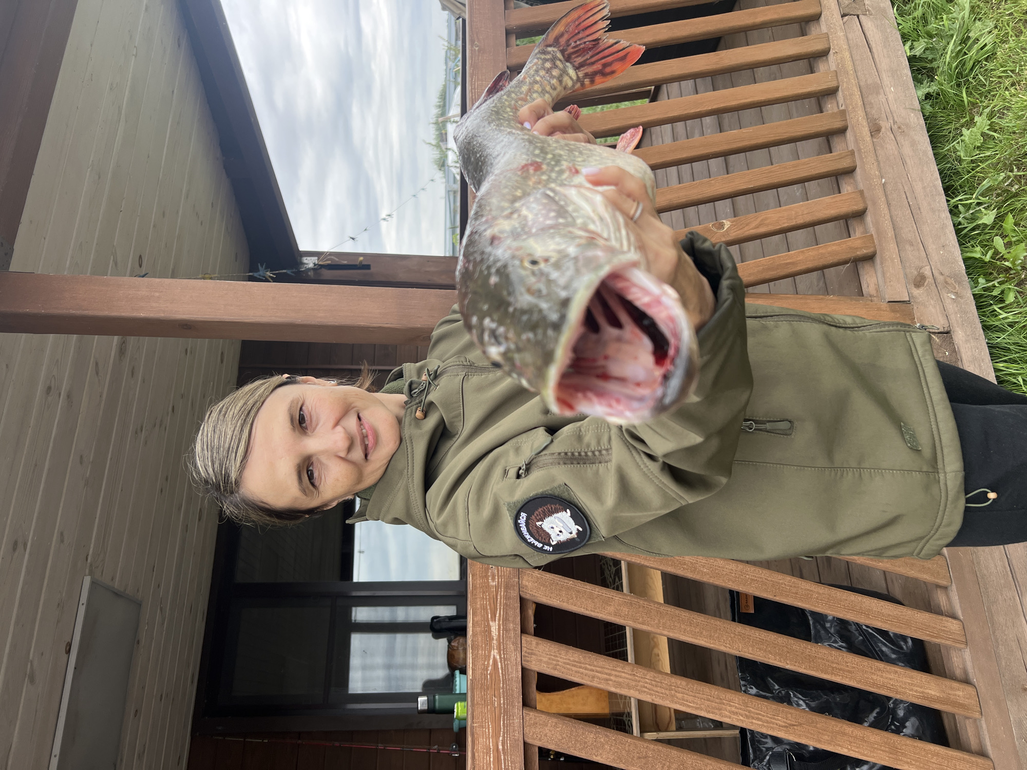 A woman stands on a wooden porch, holding a large, freshly caught fish with its mouth open. She is wearing a green hooded jacket with a patch on the left sleeve depicting a hedgehog. The background shows wooden railings, the side of a wooden building with windows, and a cloudy sky. The location is specified as Ново-Пичуго, Russia.