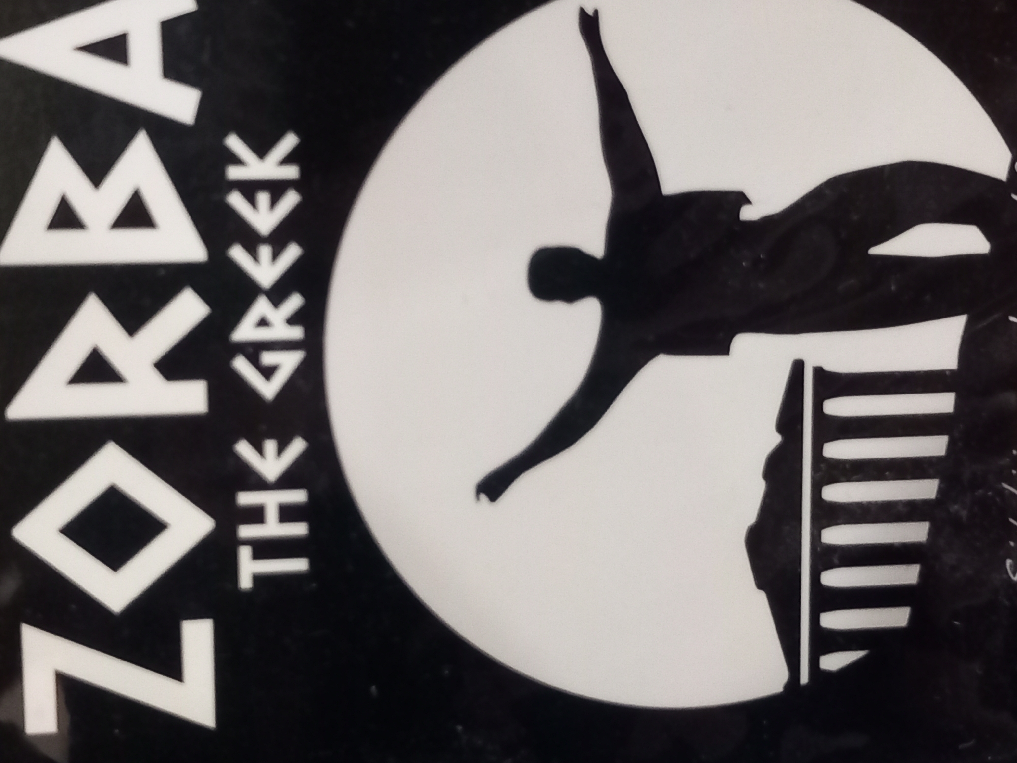 The image is a black and white graphic that appears to be a poster or album cover. The title "ZORBA THE GREEK" is displayed prominently at the top in white, block lettering. Below the title, a silhouetted figure of a man with arms outstretched is depicted within a large circle. To the left of the figure, within the circle, is a silhouette of a classical Greek building with columns. The overall style is minimalist and iconic. No city or environmental context is provided.