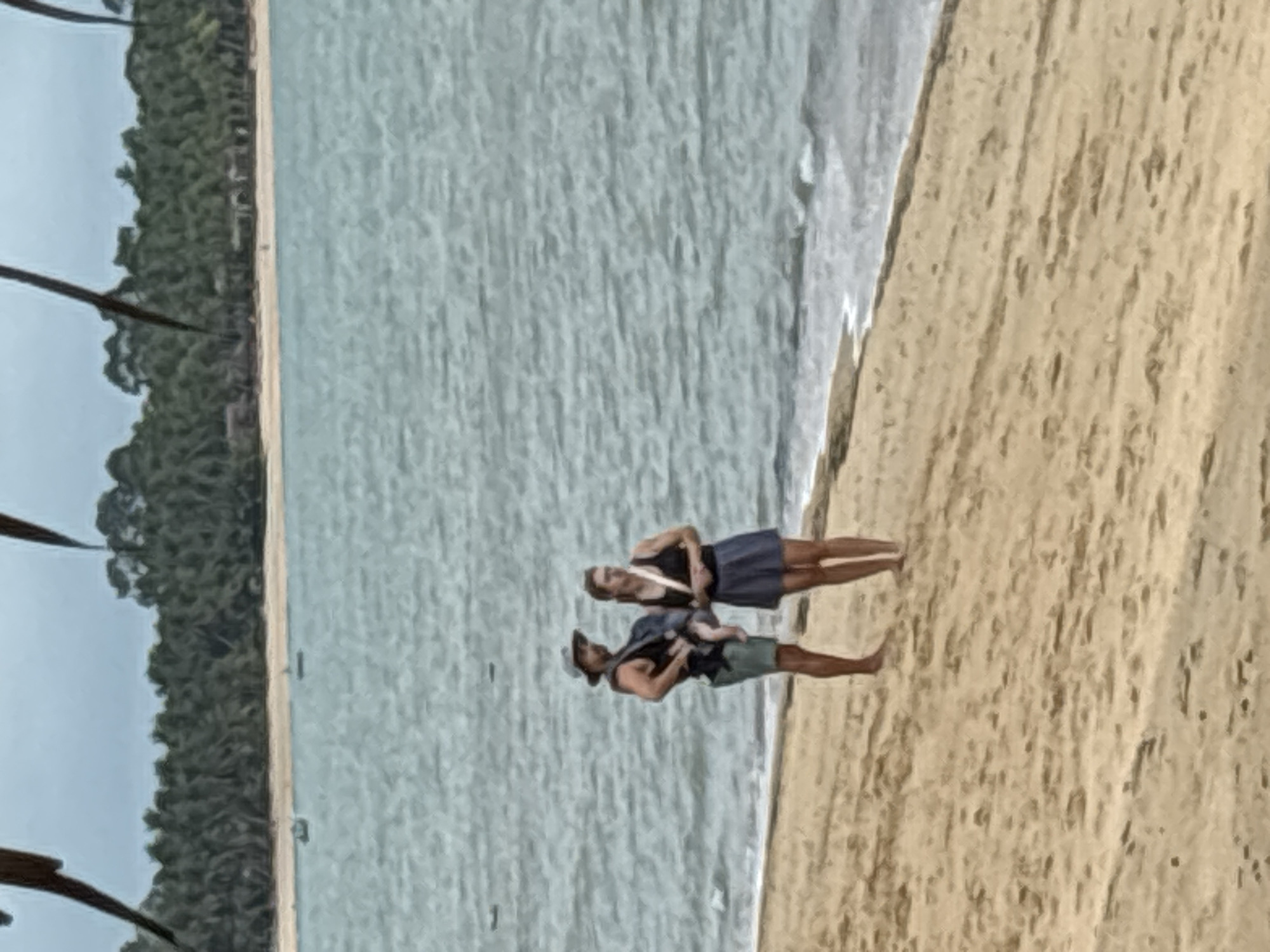 The image depicts a beach scene, likely in Baan Bo Phut, Thailand, as indicated by the tropical vegetation. A man and a woman are standing on the sandy beach, with the ocean to their left. The man is carrying a baby in a carrier on his chest. The woman is standing next to him. The beach has wet sand near the water. The ocean is slightly choppy. Some unidentified palm fronds are present in the foreground of the image, but not obscuring the subjects.