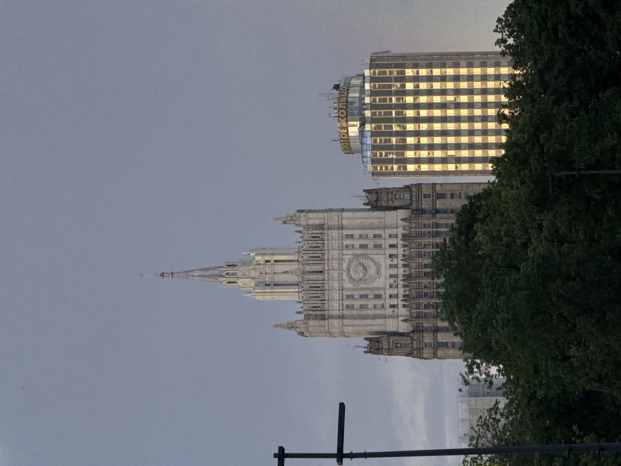 The image shows a view of the Seven Sisters skyscrapers in Moscow, Russia, against a cloudy sky. The prominent Stalinist skyscraper, the Ministry of Foreign Affairs building, is on the left, with its distinctive spire. To its right is the Golden Ring hotel building, a more modern skyscraper with a rounded top and a facade of glass windows that reflect the golden light. In the foreground, dark green trees obscure the lower portions of the buildings. A dark, cross-shaped structure, possibly a street lamp or part of a communication tower, is visible on the left edge of the frame. The overall impression is one of urban architecture and natural elements under an overcast sky, possibly during twilight.