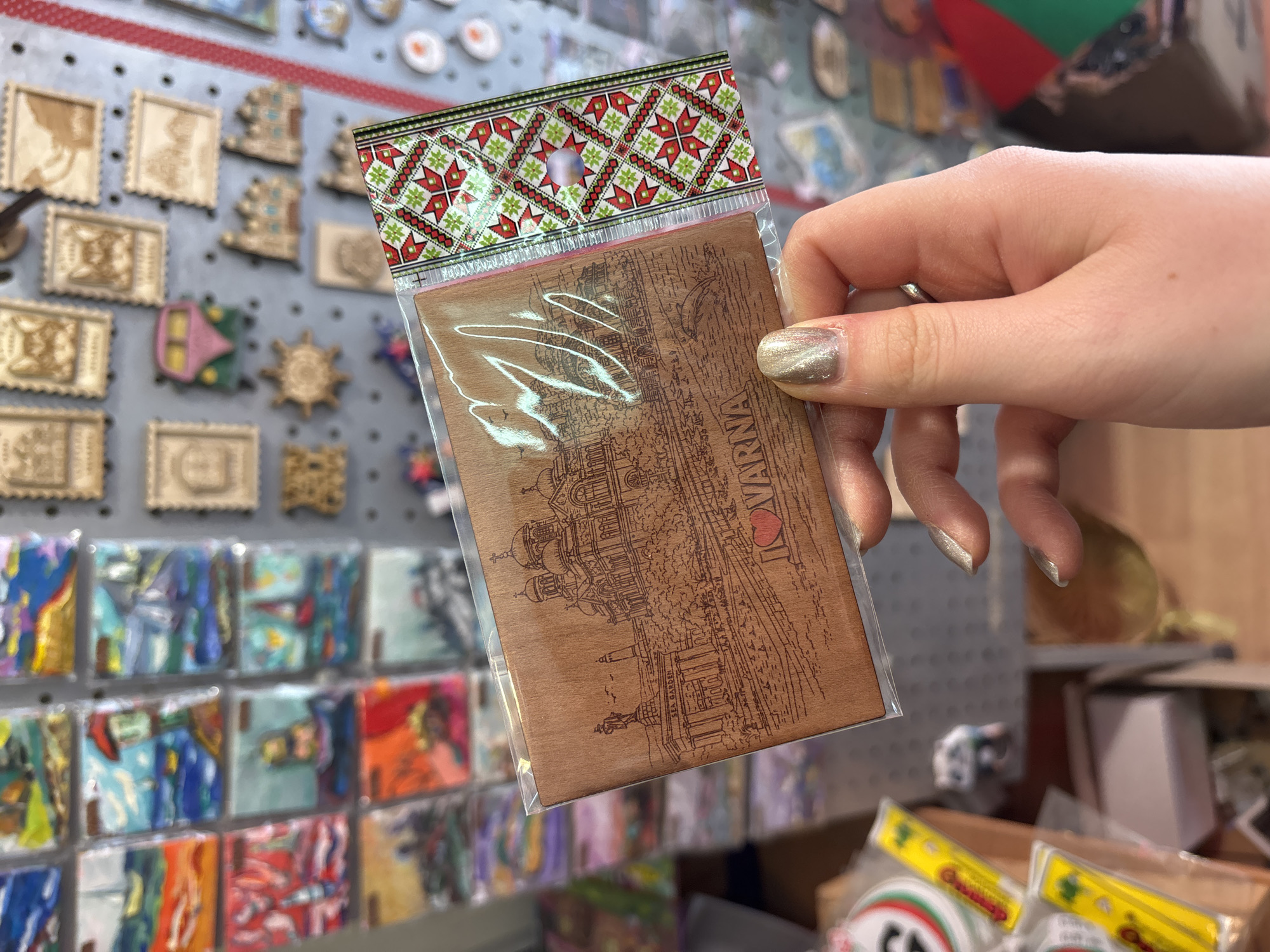 A hand holds a souvenir item, a wooden plaque depicting the cityscape of Varna, Bulgaria, encased in a clear plastic sleeve with a decorative patterned header. The artwork on the plaque shows architectural landmarks and the name "Varna" alongside a heart symbol. The background features a display wall covered with various other souvenirs.