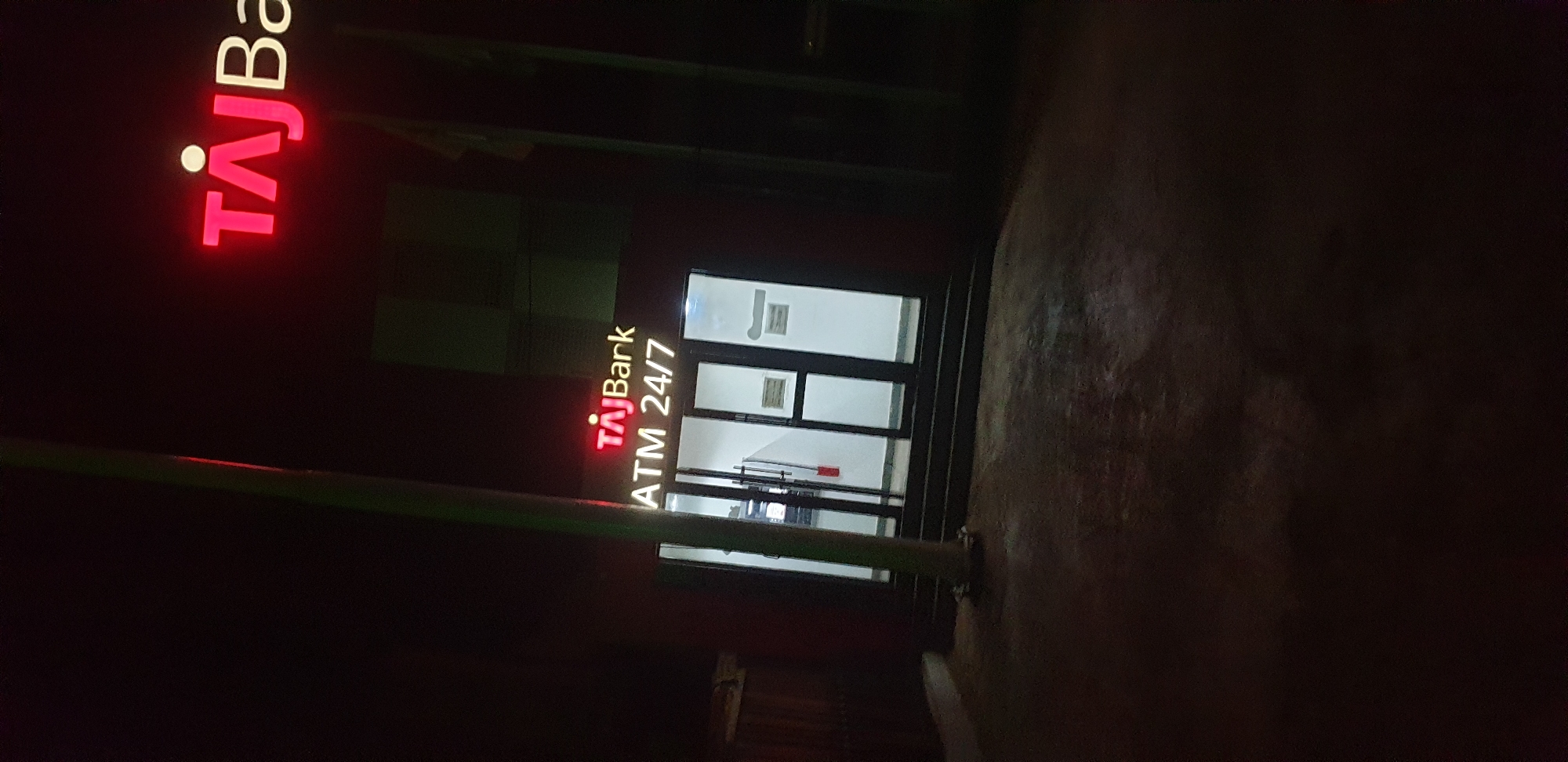 The image depicts the exterior of a TAJBank ATM facility at night, located in Sabon Tasha, Nigeria. The building features a prominent, illuminated red sign with "TAJBank" and "ATM 24/7" in white lettering below it, indicating 24-hour service. The structure itself is dark-colored with a glass entrance composed of multiple panels, accessed via a short flight of steps. Inside the brightly lit vestibule, an ATM machine is visible on the left, and a red fire extinguisher or similar object is mounted on the left wall. Several white signs or notices are affixed to the right interior wall. In the foreground, a tall, slender pole with a green hue stands vertically, partially illuminated by the bank's signage. The ground directly in front of the entrance is a dark, unadorned concrete or paved surface. To the far right, two dark flagpoles with what appear to be dark flags are faintly discernible against the minimal ambient light. No individuals are present in the scene.