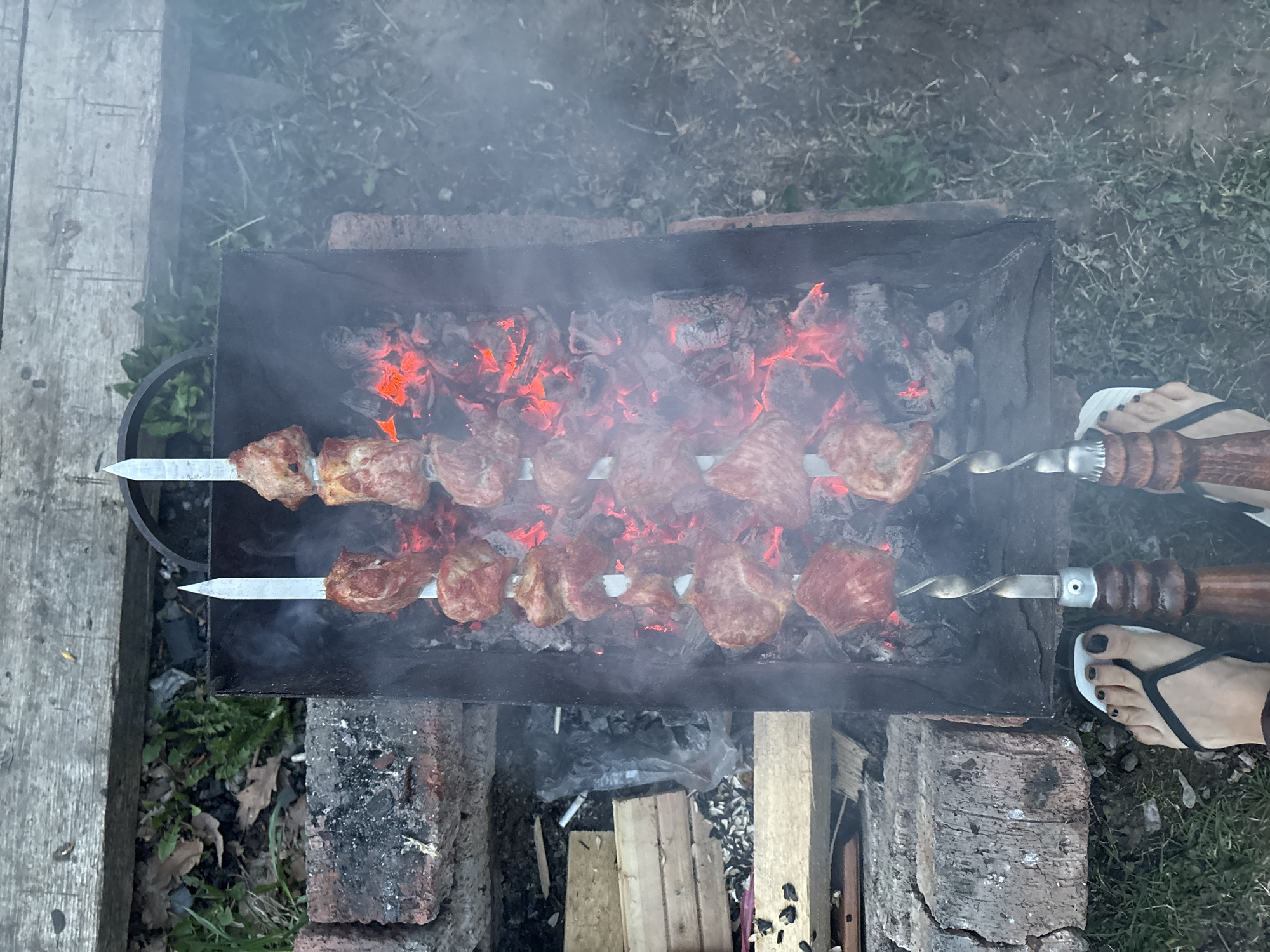 An overhead shot depicts a barbecue grill in an outdoor setting, possibly near Chaikovsky, Russia. Two metal skewers laden with meat, likely shashlik, are placed over a bed of glowing embers and charcoal within the grill. The grill appears to be made of black metal and is situated on a makeshift base constructed from bricks and wooden planks. Smoke rises from the grill, partially obscuring the background. A pair of feet wearing flip-flops are visible in the lower right corner of the frame, positioned near the grill. The surrounding environment includes grass and some form of vegetation.