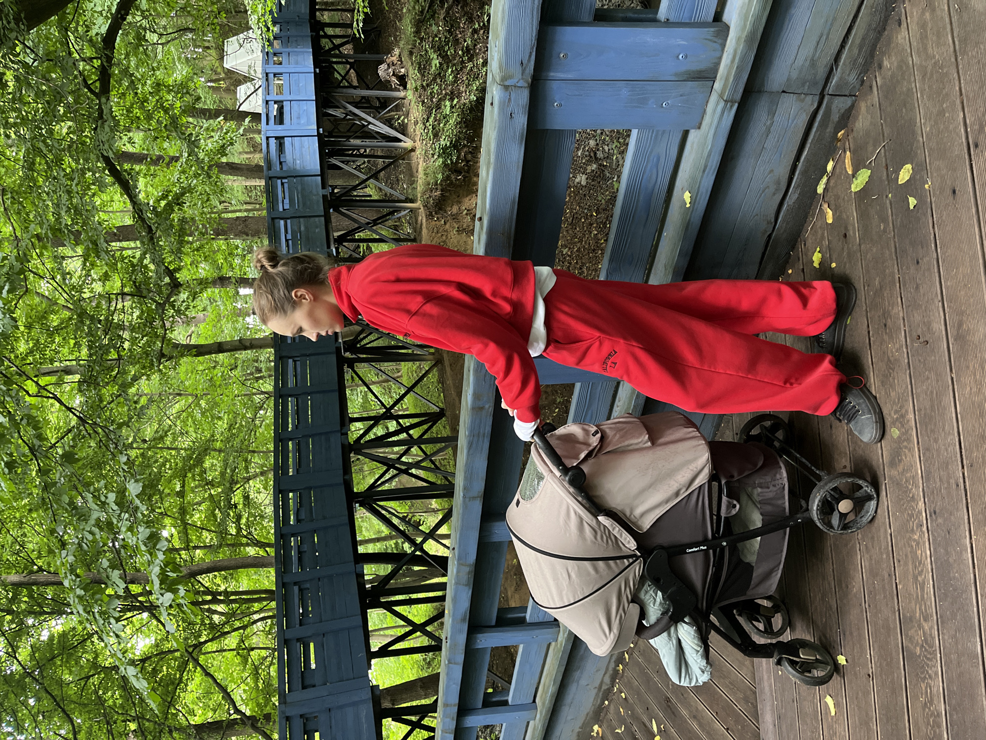 In Pionersky, Russia, a young woman in a red tracksuit pushes a beige stroller across a wooden boardwalk. The boardwalk is bordered by a blue wooden railing, and behind it, a blue wooden structure resembling a bridge or walkway extends into a forest. The forest is dense with green foliage, and the boardwalk is littered with fallen leaves. The woman is facing away from the camera, with her hair tied up in a bun. The stroller is partially covered, obscuring its contents. The scene appears to be an outdoor recreational area within a wooded environment.