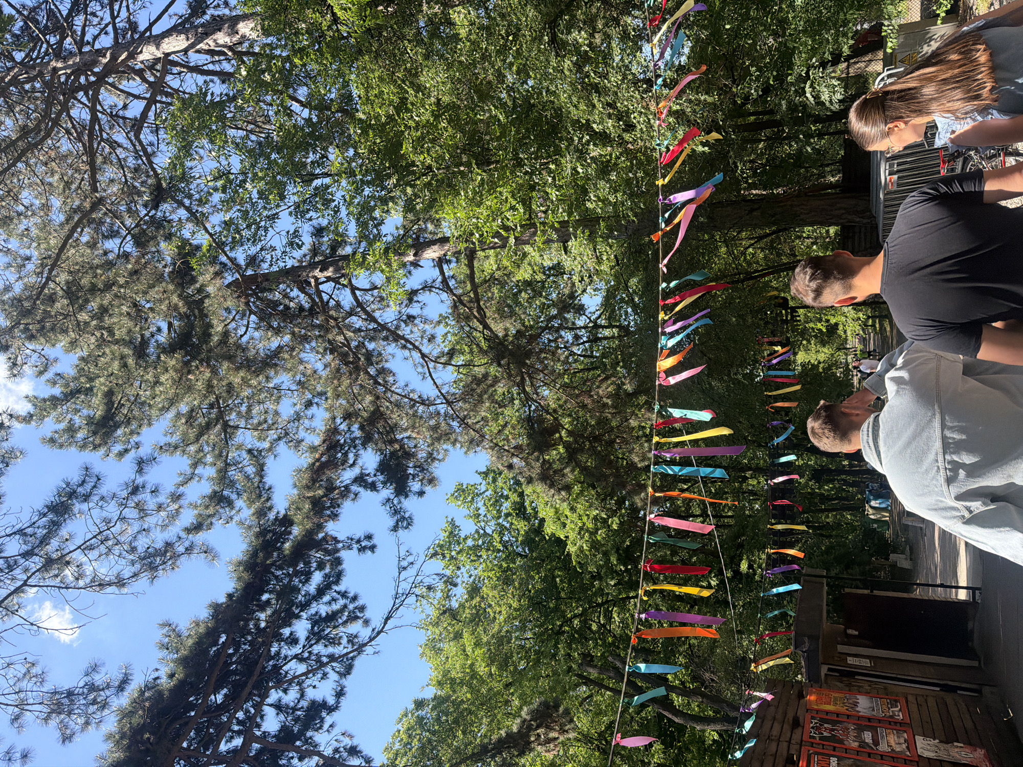 The image shows a park in Sofia, Bulgaria, under a bright, daytime sky. Trees with green foliage and tall pine trees frame the scene, partially obscuring the blue sky. Strands of colorful ribbons are strung between the trees, adding a festive touch. Three people are visible, with their backs to the camera; one is wearing a light blue denim shirt, another is wearing a black t-shirt, and the third is wearing a light blue dress. The ribbons are suspended above a path or walkway, and a building or structure with posters on its exterior is partially visible.