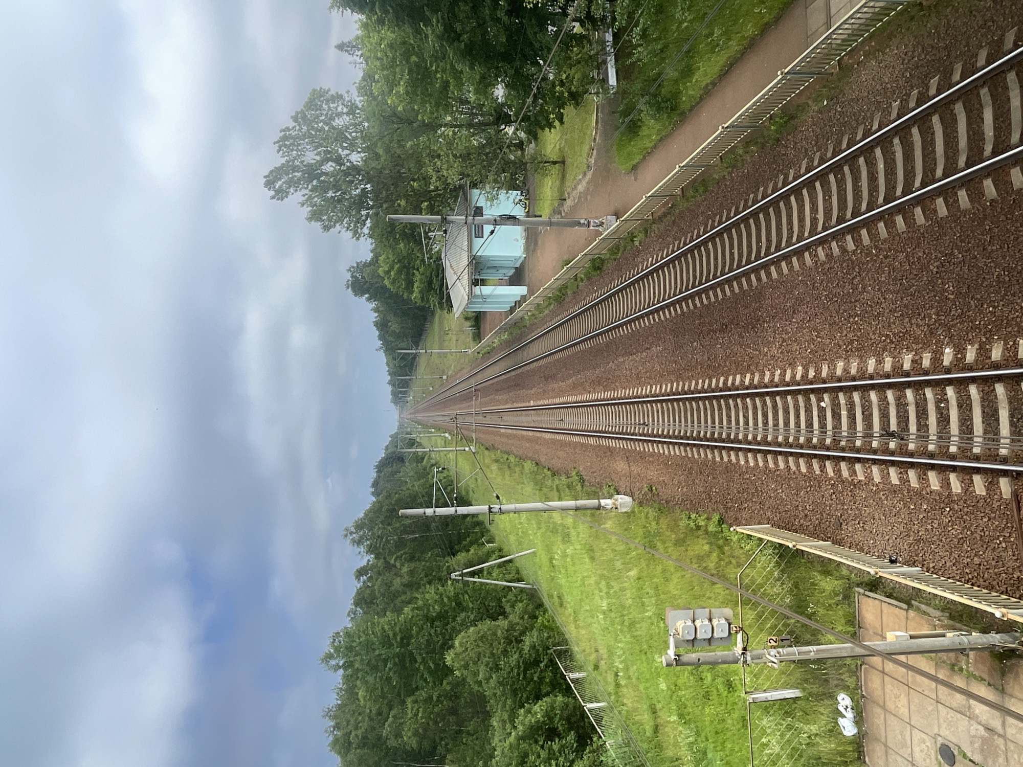 The image shows a railway line running through a green, forested area. The tracks are in the foreground, laid on a bed of brown ballast, with white sleepers visible. To the left of the tracks, there is a grassy verge and a dense line of trees. In the distance, a small, light blue building with a slanted roof is situated beside the tracks, possibly a small station or maintenance shed. Overhead power lines stretch along the railway. The sky is overcast with gray clouds, suggesting a cool or possibly rainy day. The image is oriented vertically, with the railway receding into the distance towards the top. There is no visible activity or people. The scene conveys a sense of quiet solitude and rural transportation. There is some text on a sign on a pole near the foreground, which appears to be the number "2". The location context is Solnechnoye, Russia.