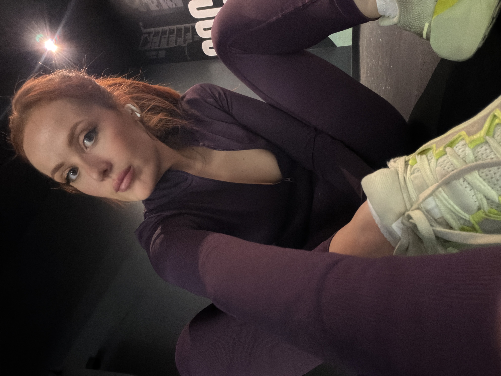 A low-angle selfie depicts a woman with fair skin and auburn hair pulled back into a ponytail. She wears an in-ear headphone in her left ear. She is dressed in a plum-colored athletic outfit, consisting of a long-sleeved jacket that is slightly open at the top. Her legs are crossed, and she is wearing white athletic shoes with neon green accents. The background is dark and indistinct, but a bright light is visible in the upper left corner of the frame. Part of a wall with white lettering and what appears to be a metal structure is seen in the background. The location cannot be determined from this image.
