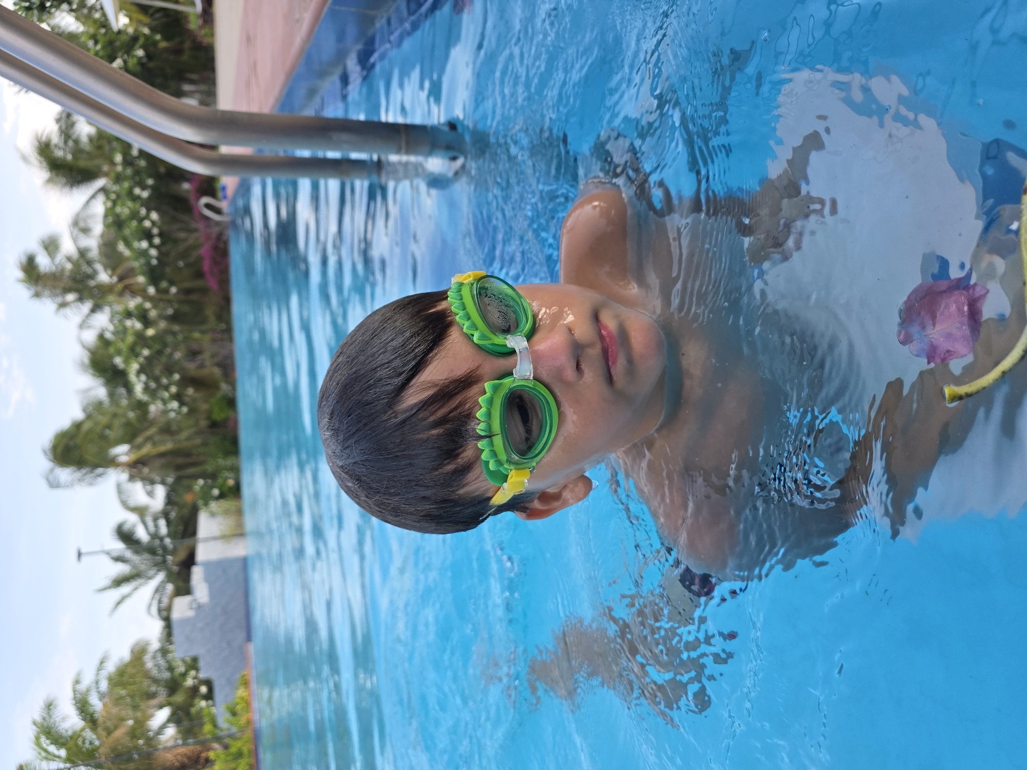 A child wearing green goggles is in a swimming pool. The pool is surrounded by vegetation, and a metal railing is visible. The location is Phu Thuy Ward, Vietnam.