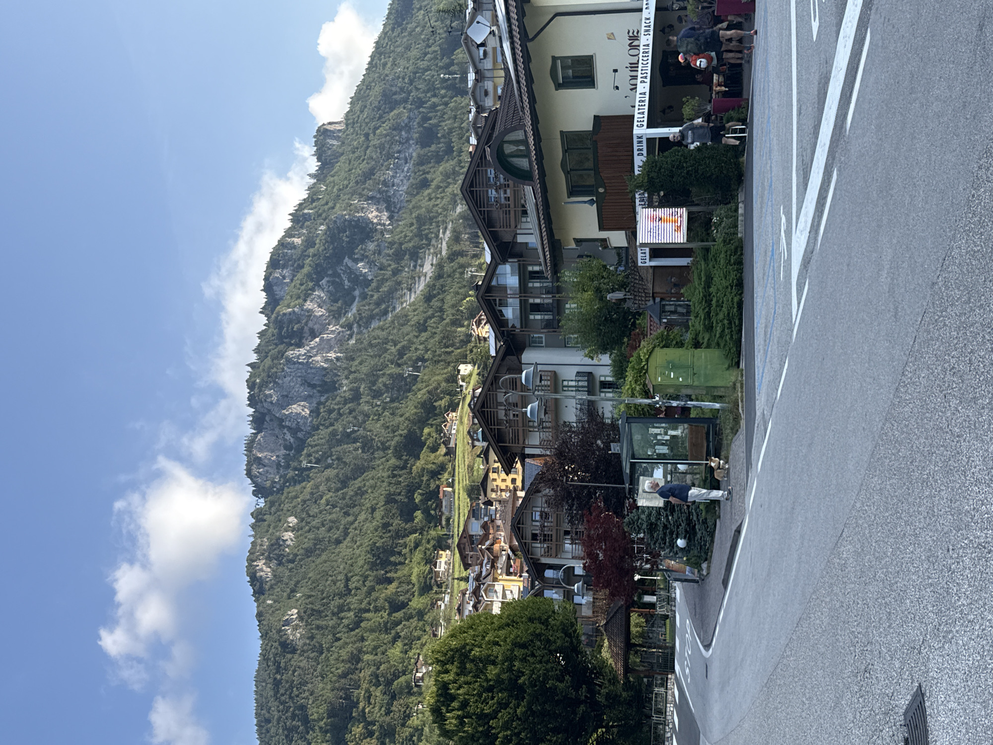 The image depicts a street scene in the town of Molveno, Italy, with a steep, forested mountain rising in the background under a partly cloudy sky. In the foreground, a paved road curves to the left, marked with white lines and a "STOP" sign. On the right side of the road, a building with the signage "GELATERIA - PASTICCEREIA - SNACK" is visible, with outdoor seating and patrons. Several people are present: a man in a blue shirt and light-colored pants stands with a small dog near a bus stop shelter on the sidewalk. Further down the street, a man in a gray t-shirt and black shorts stands by a digital display screen, and two individuals, one with a backpack and the other wearing a pink hat, are walking away from the camera. The architecture of the buildings is characteristic of a European mountain town, with wooden balconies and pitched roofs. The overall impression is a peaceful, late afternoon in a scenic Italian alpine village.