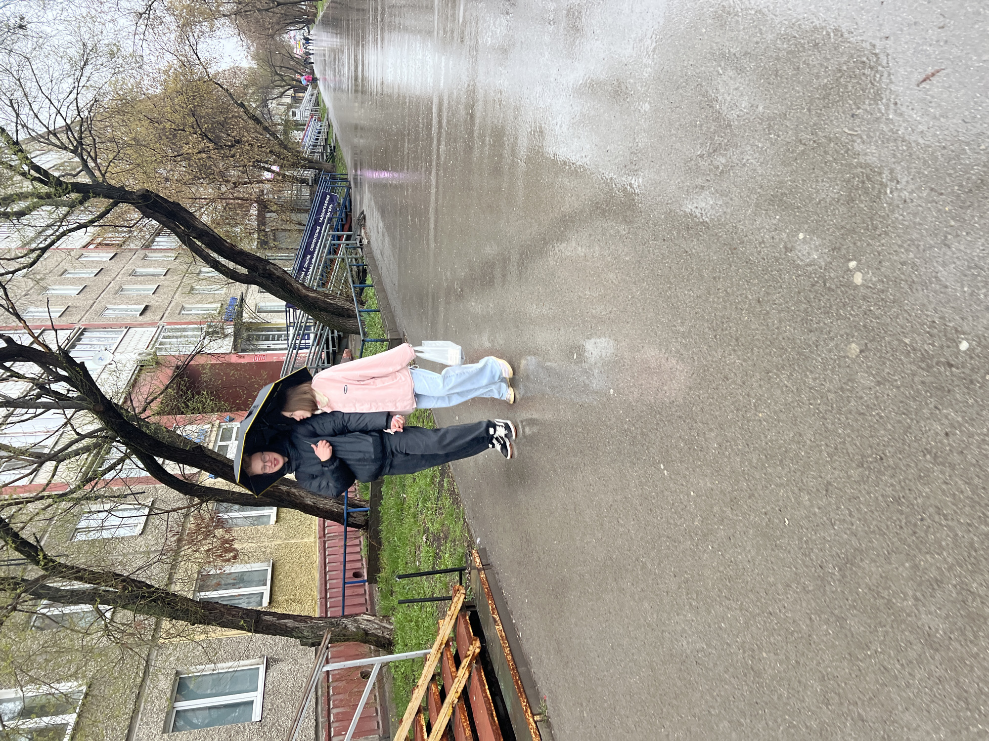 In Perm, Russia, two individuals walk under an umbrella on a wet, paved street. One person wears a black jacket and pants, and the other wears a light pink jacket and light blue jeans. The area includes multi-story apartment buildings and bare trees. An old metal fence is along the edge of the sidewalk.