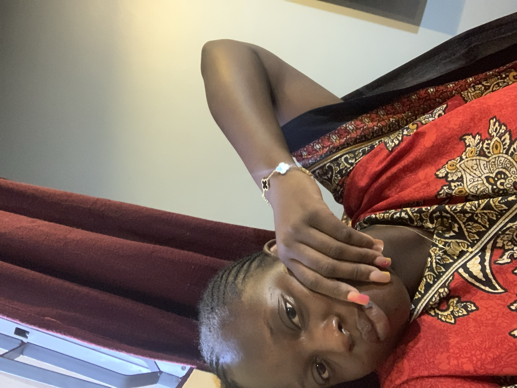 A young Black woman is shown in a close-up, selfie-style shot. She has her right hand on her face, partially obscuring her cheek and mouth. Her hair is styled in braids. She is wearing a red and black patterned dress and a bracelet on her left wrist. The background shows a burgundy curtain to the left and a light-colored wall to the right. The lighting appears to be artificial, casting a warm glow on the wall. The image does not provide any specific details to determine the location in the city.