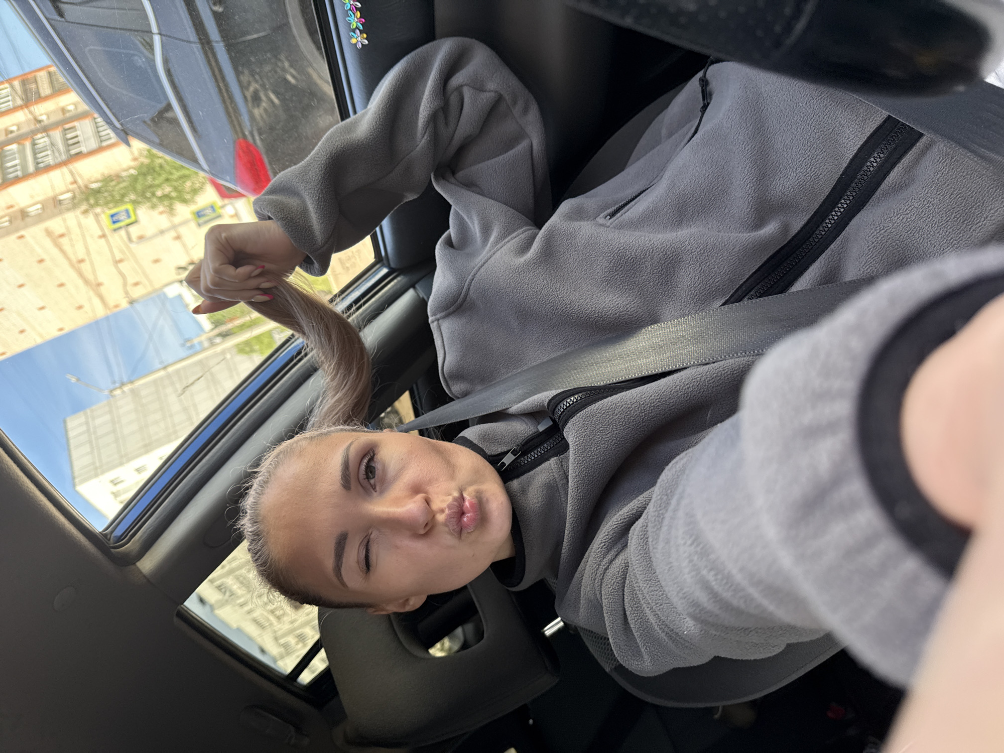 A fair-skinned female sits in a car in Chaikovsky, Russia. She wears a grey zip-up fleece jacket with a black zipper. A seatbelt crosses her chest. She holds a long, light brown ponytail in her left hand, visible near the car window. The woman winks and puckers her lips toward the camera. Through the open sunroof, a building and blue sky are visible. Car interior details include the headrest and dashboard. A glimpse of a street sign and more buildings are seen through the side window. Decorative flower stickers are on the car's interior.