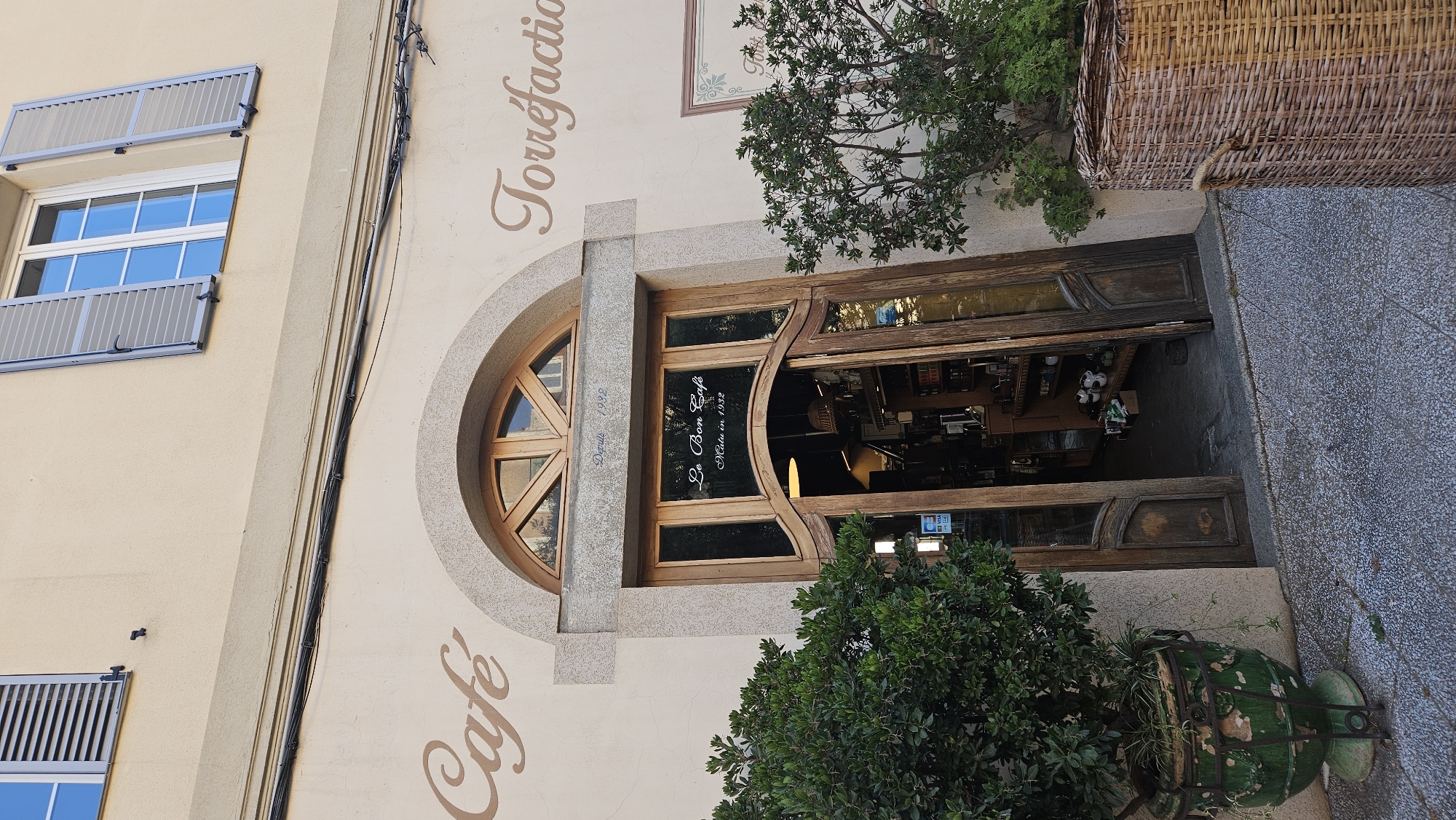 The image shows the exterior of a shop in L'Île-Rousse, France, identified by the text "Le Bon Café" and "Malu in 1932" on the glass door. The shop has a large arched wooden door that is partially open, revealing shelves stocked with items inside. The facade of the building is cream-colored, with a large arched window above the entrance. To the left of the entrance, the word "Café" is written in cursive. To the right, the word "Torréfaction" is also written in cursive. Two potted green plants are placed outside the shop, one in a green weathered pot and the other in a large woven basket. Above the entrance, a small sign reads "Depuis 1932". Two windows with grey shutters are visible on the upper level of the building. The ground in front of the shop is paved with stone.