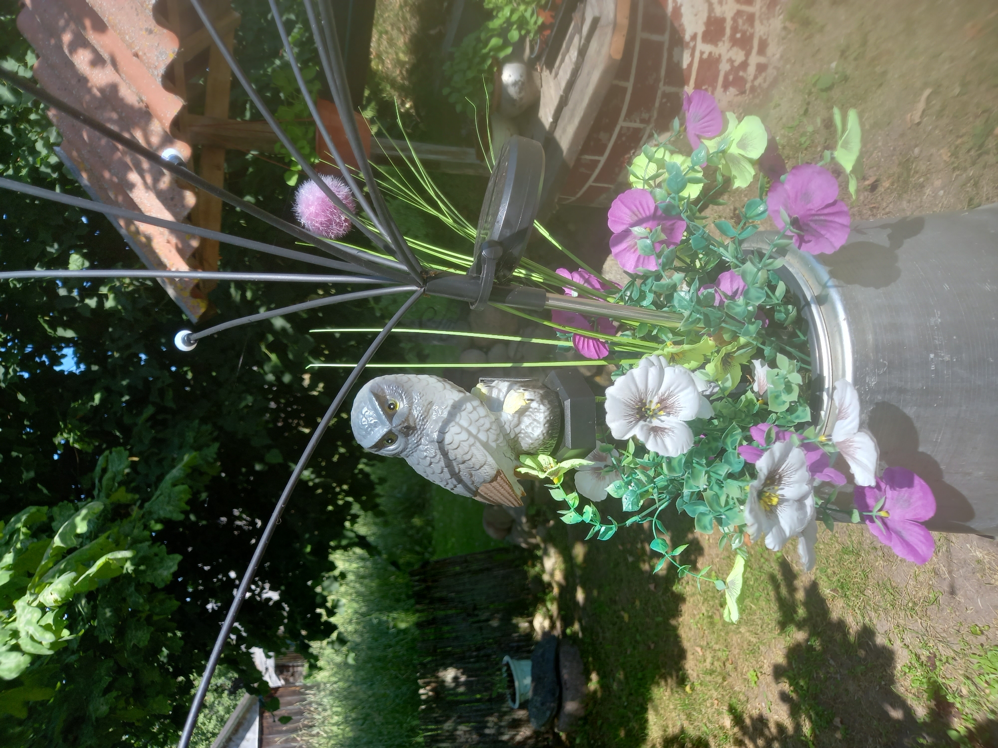 The image shows a close-up of decorative items in an outdoor setting, likely a garden. The main subjects are two owl figurines, one larger and more detailed than the other, positioned in front of artificial flowers. The larger owl is grey with distinct feather patterns and yellow eyes, and it appears to be perched on something. Surrounding the owls are artificial flowers in shades of purple, light yellow, and white, with green leaves and stems. These flowers are arranged in a metallic container, possibly a bucket or planter.
Behind the decorative elements, glimpses of the surroundings can be seen. There is a reddish-brown tiled roof on the left, and a brick structure in the background to the right. Lush green foliage from trees or bushes forms a backdrop. The ground is covered in grass and some patches of dry earth. The lighting suggests it is daytime, with sunlight casting shadows, indicating a sunny day.
The overall impression is of a decorated outdoor space, possibly a garden or patio, with the owl figures and artificial flowers serving as ornamentation. There is no activity or event taking place, and no people are visible. The scene is static and focuses on the decorative elements and their immediate surroundings. No visible text is present in the image. The location context of Adutiškis, Lithuania, suggests a possibly rural or suburban setting.