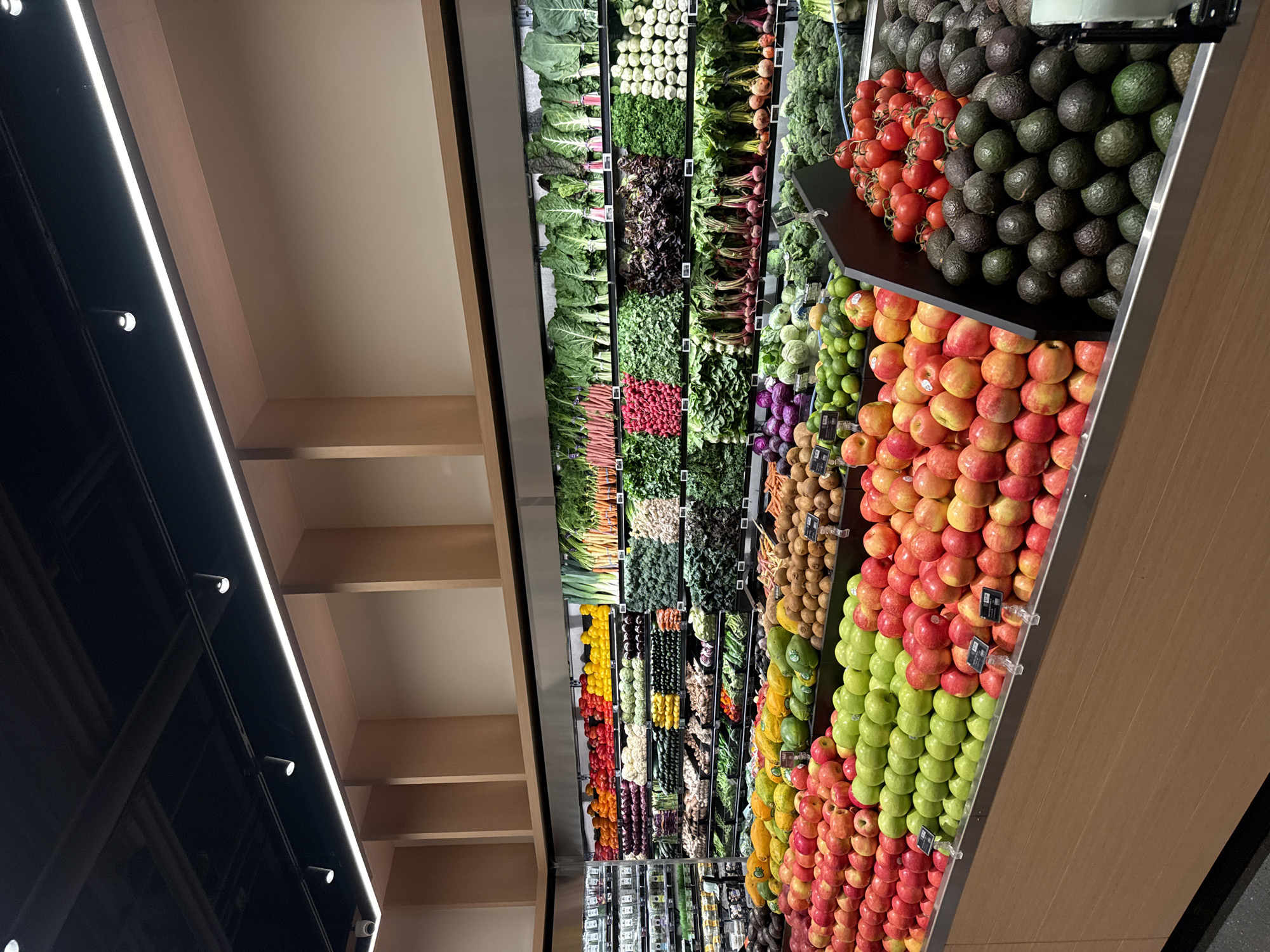 The image shows a produce display at a grocery store, likely in West Hollywood, United States of America. The display is brightly lit and filled with a wide variety of fruits and vegetables arranged in neat rows. The produce includes leafy greens, tomatoes, avocados, apples (both green and red), root vegetables, citrus fruits, and other fresh items. The presentation is meticulous, with produce stacked high and arranged for visual appeal. The shelving system appears modern, with wood-like panels. Price tags are visible on some of the items. The lighting suggests a focus on highlighting the freshness and vibrancy of the produce.