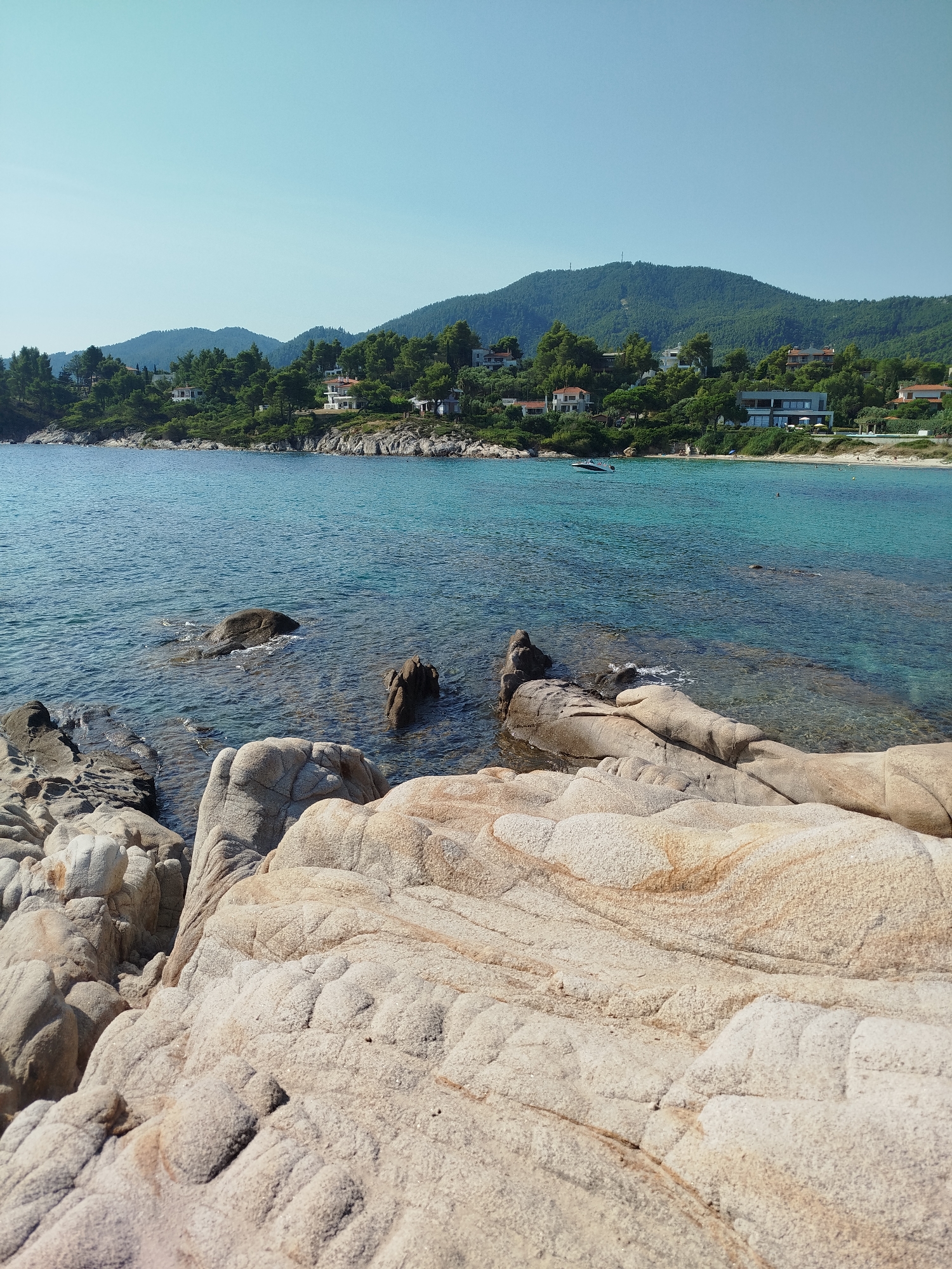 The image depicts a scenic view of a rocky coastline in Sithonia Municipal Unit, Greece. The foreground is dominated by large, light-colored rocks with weathered textures. Beyond the rocks, the clear turquoise water of the sea gently laps the shore. In the middle ground, more submerged rocks are visible beneath the surface. The background features a verdant, tree-covered hillside dotted with houses and buildings. The sky is a clear, pale blue, indicating a sunny day. A small white boat is visible on the water near the shore. The overall scene conveys a sense of tranquility and natural beauty characteristic of a coastal Mediterranean landscape.