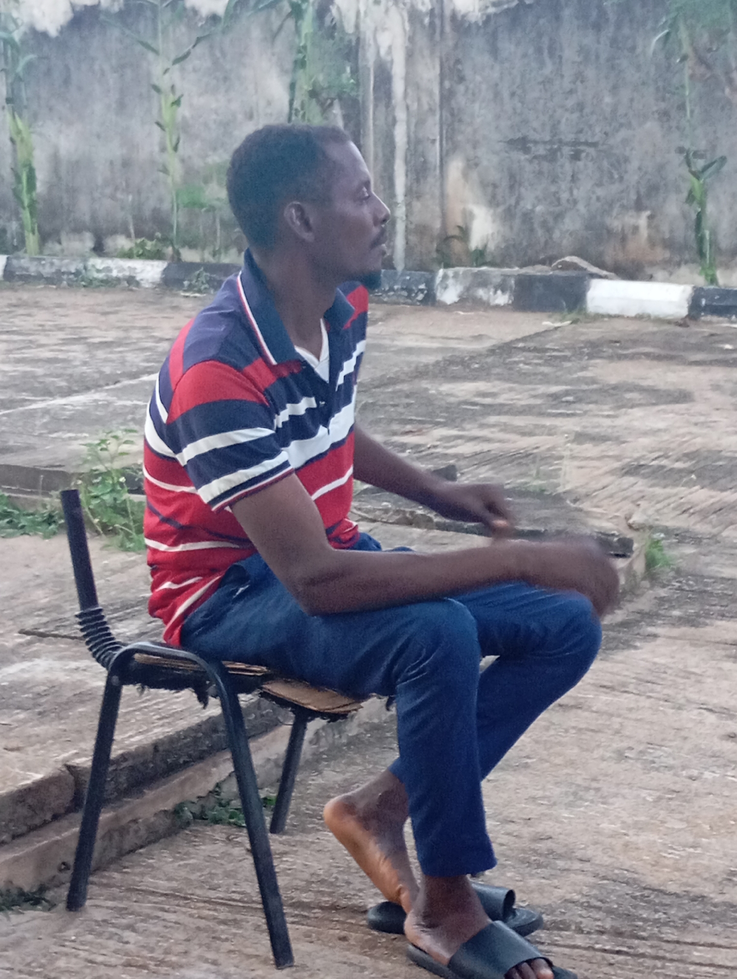 In Shendam, Nigeria, a man is seated on a black metal chair with a worn seat. He is wearing a red, blue, and white striped polo shirt and dark blue trousers. The man is looking to his right, with his right hand resting on his lap. His left arm is bent, and his left hand appears to be holding something thin and dark. His feet are bare except for black sandals. The chair is positioned on a paved outdoor surface with a textured, greyish pattern. In the background, a concrete wall is visible, with some greenery and tall plants growing near it. A black and white striped border runs along the base of the wall, separating it from the paved area. The lighting suggests it may be overcast or late in the day.
