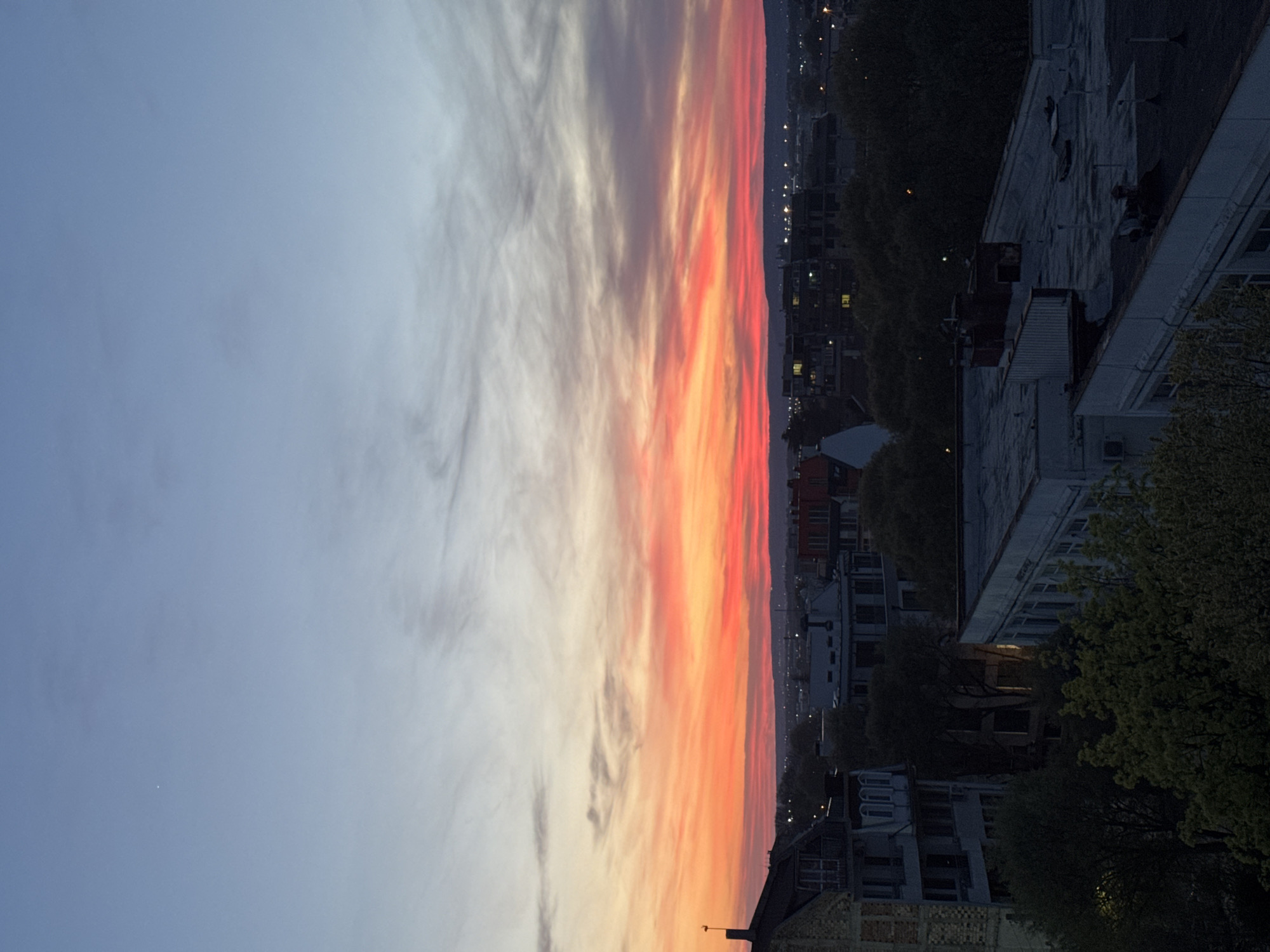 The image shows an urban landscape at dusk in Varna, Bulgaria. A striking sunset dominates the sky, with a gradient of colors transitioning from a soft blue to vibrant oranges and reds. Wispy clouds are visible in the sky. Below the sky, buildings and trees form the cityscape. The architecture suggests a mix of residential and commercial structures. The picture is rotated ninety degrees clockwise.