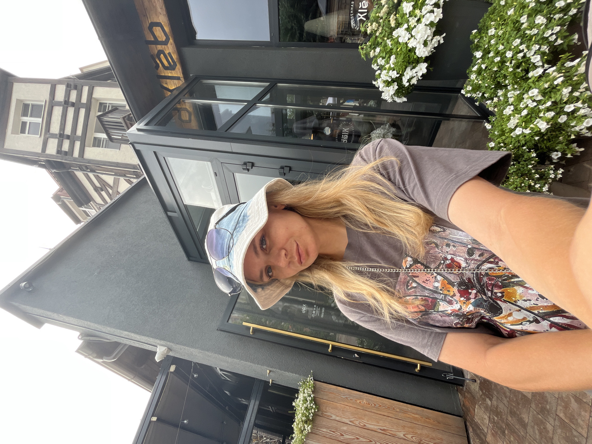 A blonde woman with fair skin, wearing a denim bucket hat, sunglasses, and a grey t-shirt with a colorful graphic print, poses for a selfie in front of a building. She is holding the camera in her right hand, extending her arm out. The building appears to be a commercial establishment with dark grey walls and large glass windows. A wooden planter box with white flowers is visible to her left, and another planter box filled with white flowers is to her right, partially obscuring the building's facade. In the background, a distinct building with a half-timbered architectural style is visible. The sign on the building's glass door, "XieXie," is an indicator that this location is likely the XieXie restaurant on Moskovskaya Street in Zelenogradsk, Russia, as this restaurant is known for its facade and the depicted building style. The overall lighting suggests it is daytime.