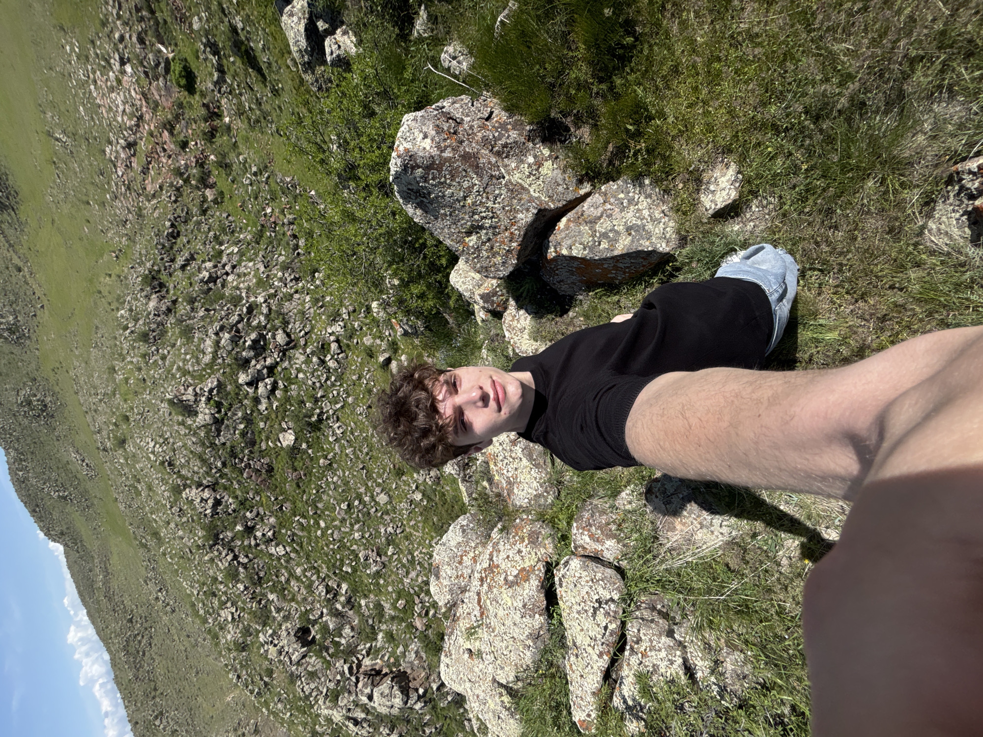 A photograph taken in Tlik, Armenia, shows a young man in a black shirt and blue jeans lying amongst lichen-covered rocks and grass on a mountainside. The landscape includes other rocks, grass, and sparse vegetation. A patch of blue sky is visible in the background. The man is facing the camera and his arm is extended towards it, indicating a self-portrait.